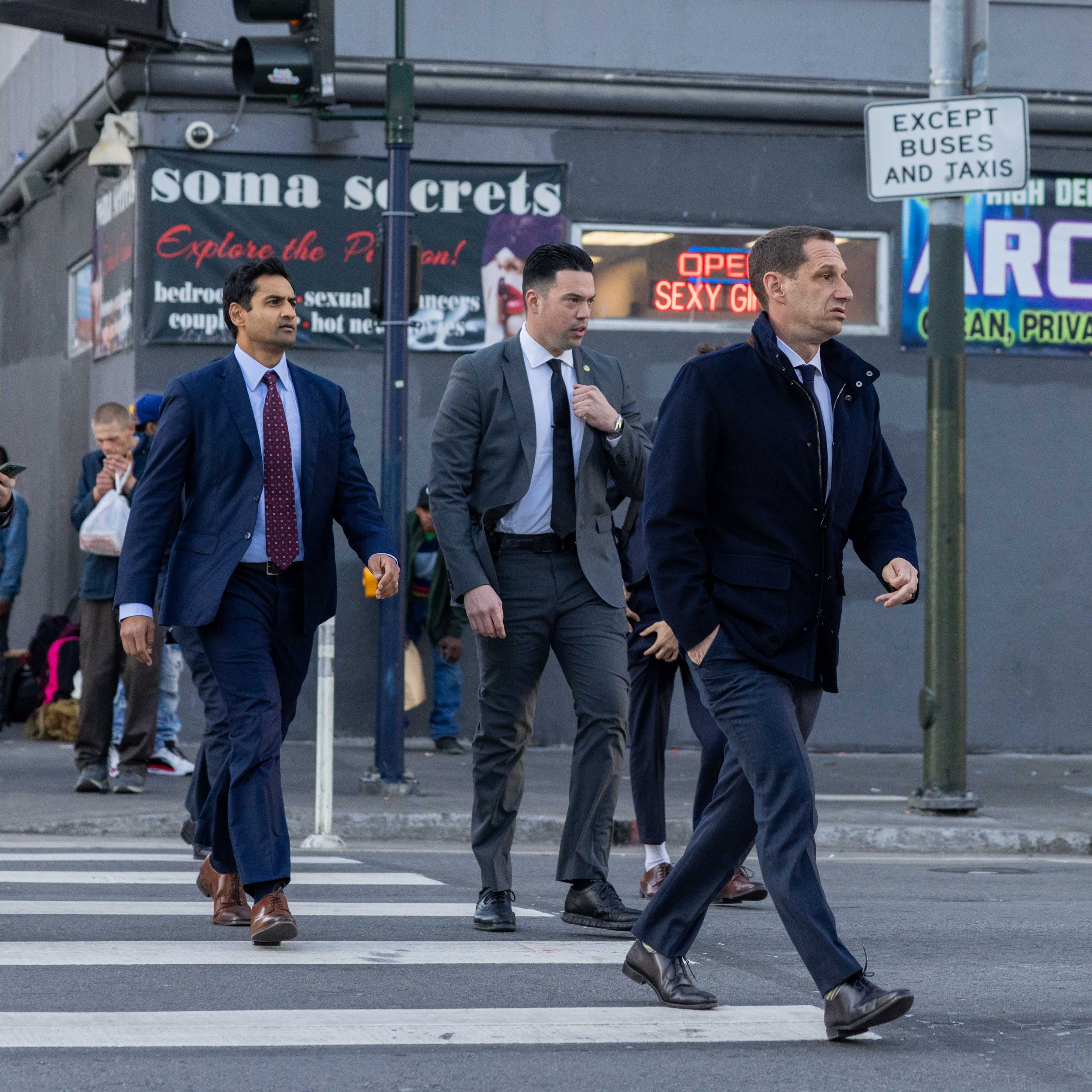 Four men in suits cross a city street, with storefronts and signs, including &quot;ARCADE,&quot; in the background. They appear focused and are walking briskly.