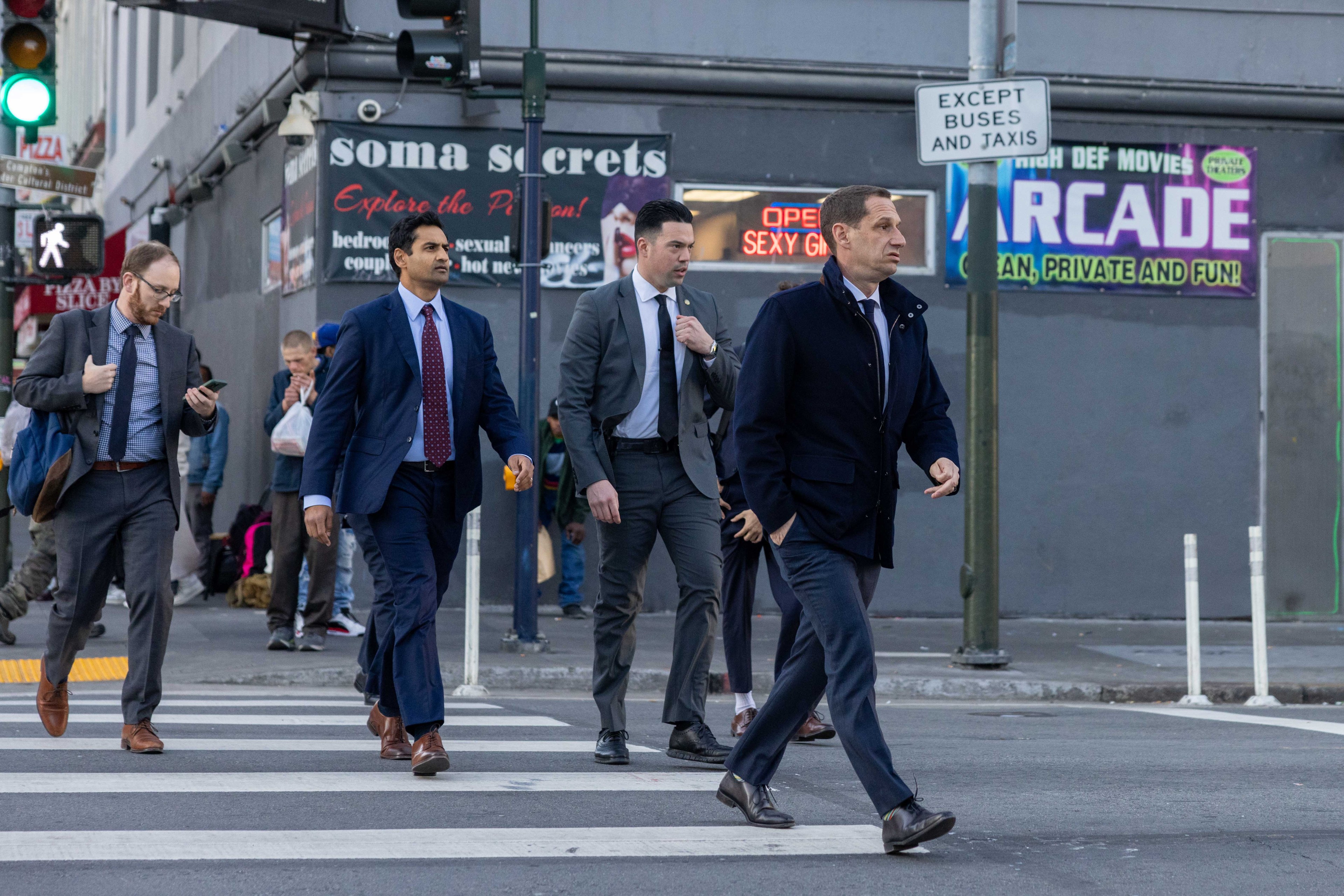 Four men in suits cross a city street, with storefronts and signs, including "ARCADE," in the background. They appear focused and are walking briskly.