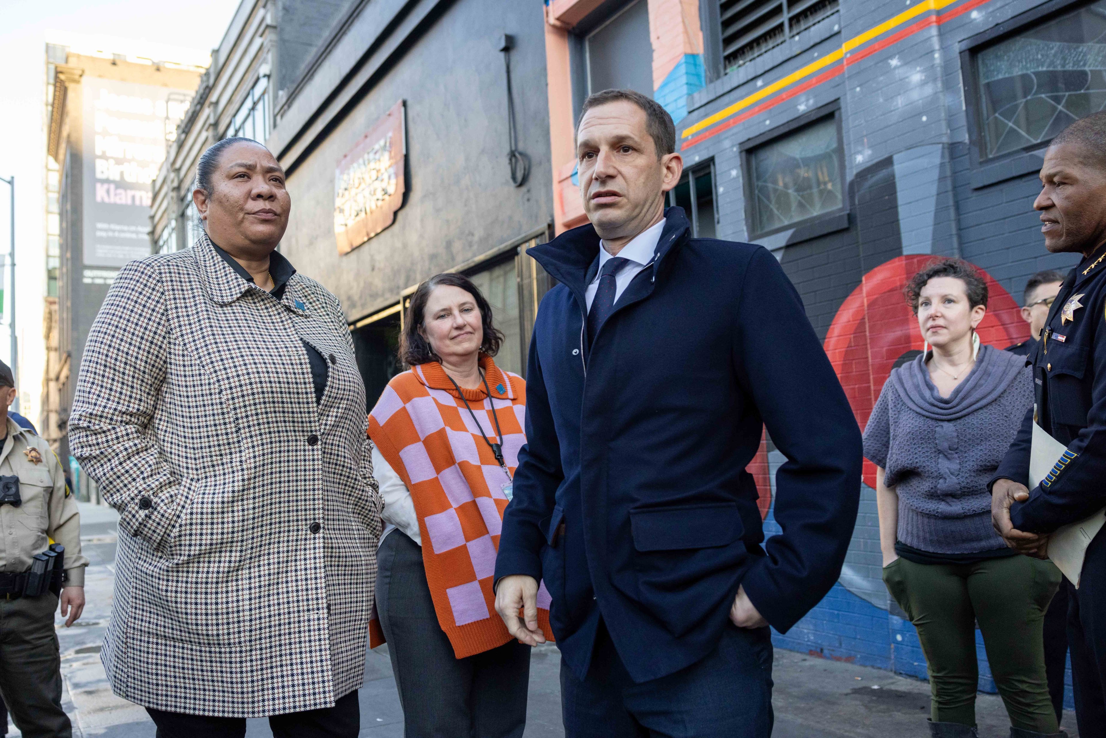 A group of people, including a woman in a checked coat and a man in a dark jacket, stand outside a colorful building with painted murals. They appear engaged.