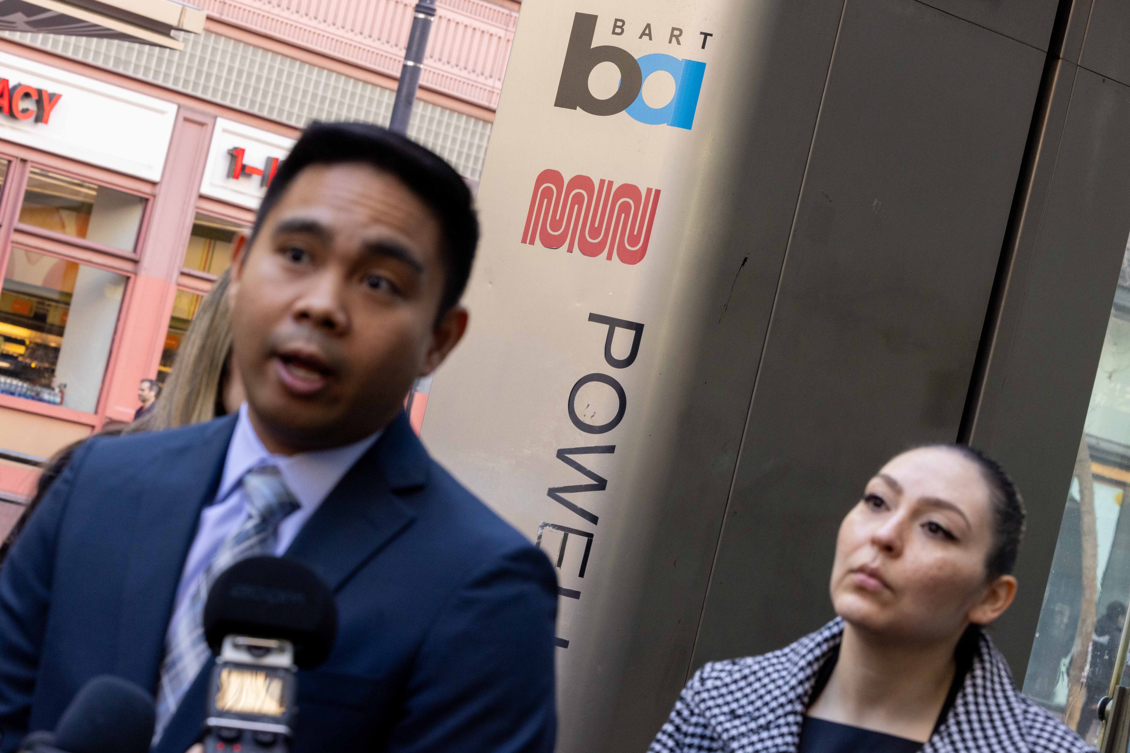 A man and woman are speaking in front of microphones. The background shows a transit station with BART and Muni logos and the word &quot;Powell&quot; visible.