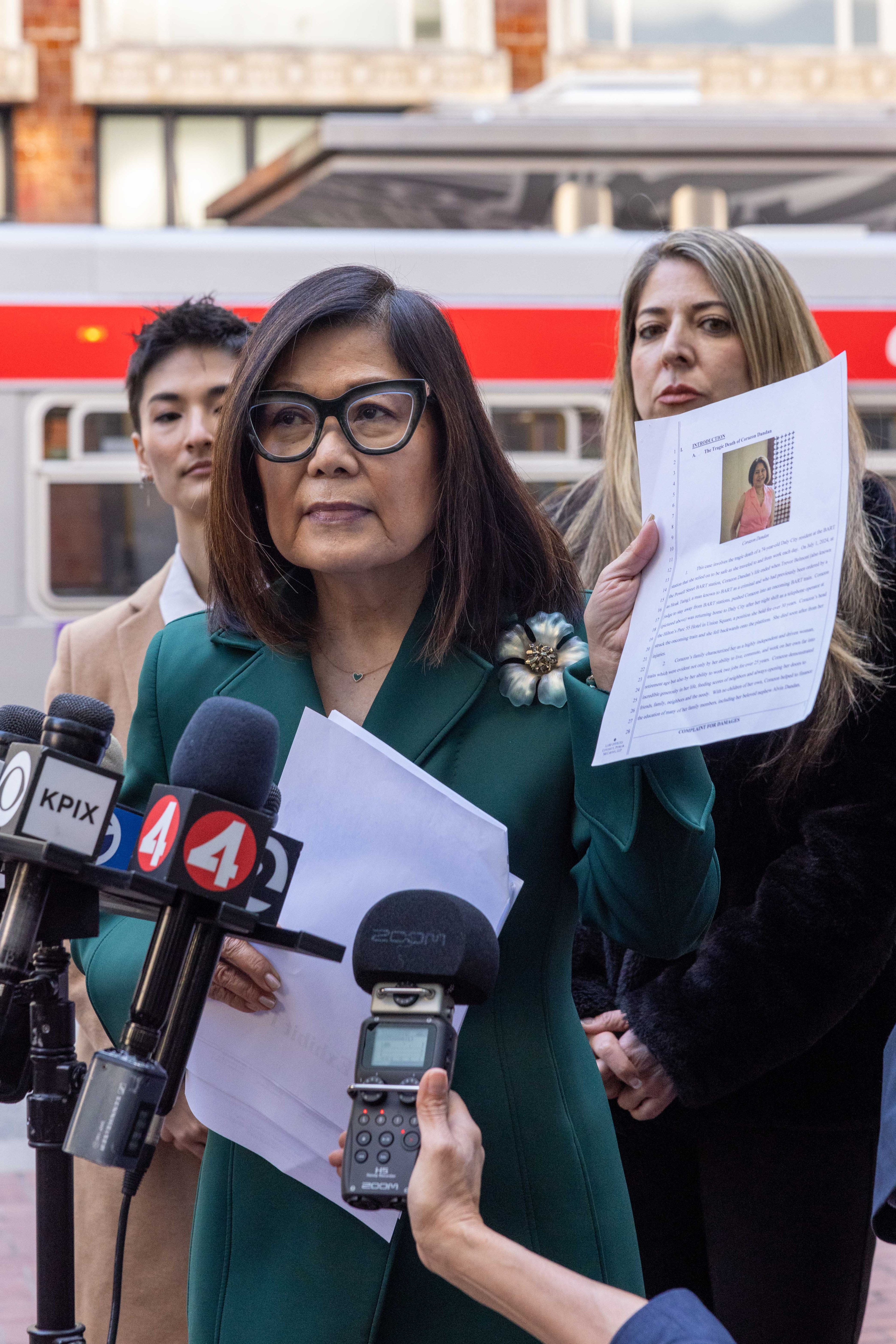 A woman in glasses addresses reporters, holding documents with a photo. Microphones from various news outlets are directed toward her.