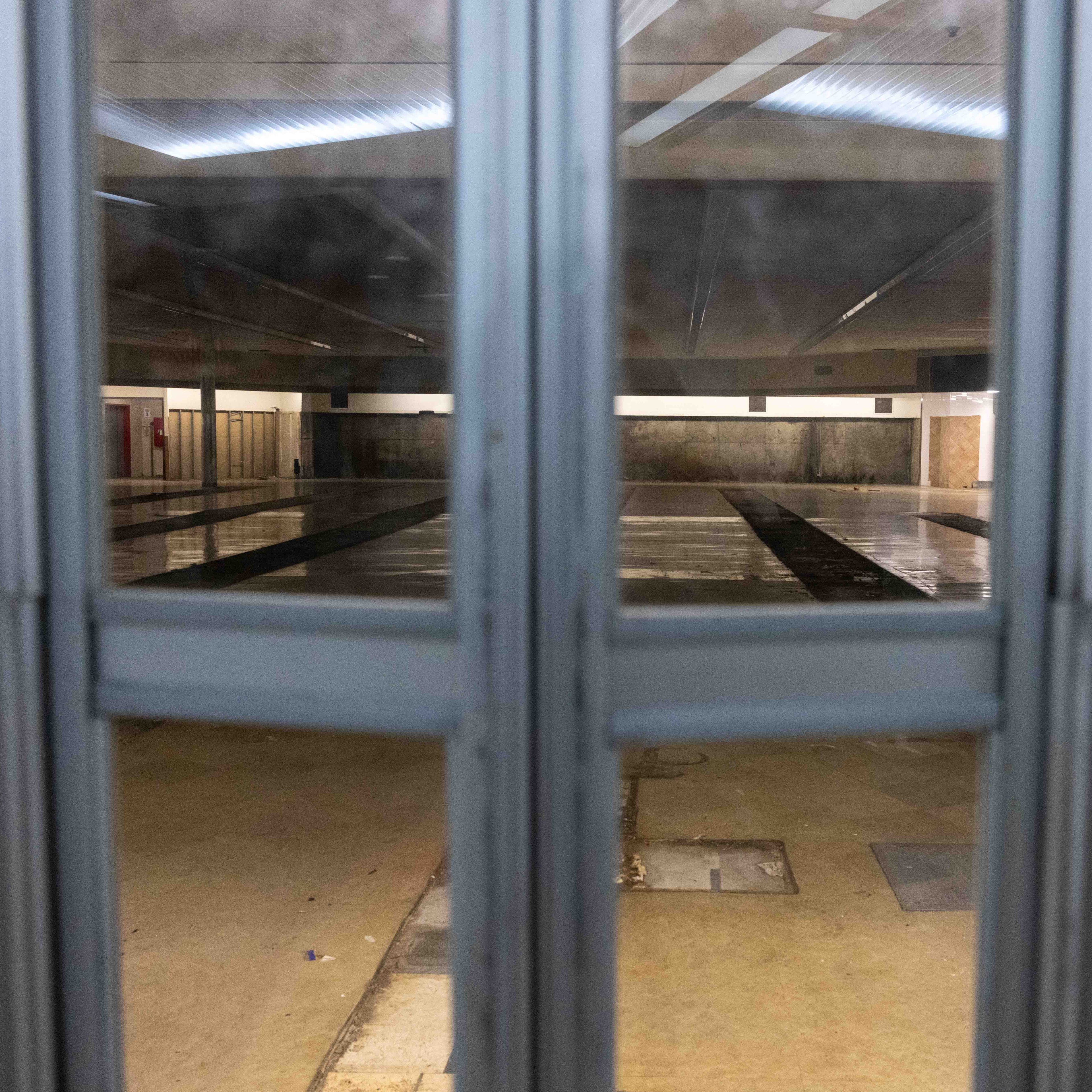 The image shows an empty indoor swimming pool, viewed through a set of glass doors, with lanes visible on the pool floor and overhead lighting.