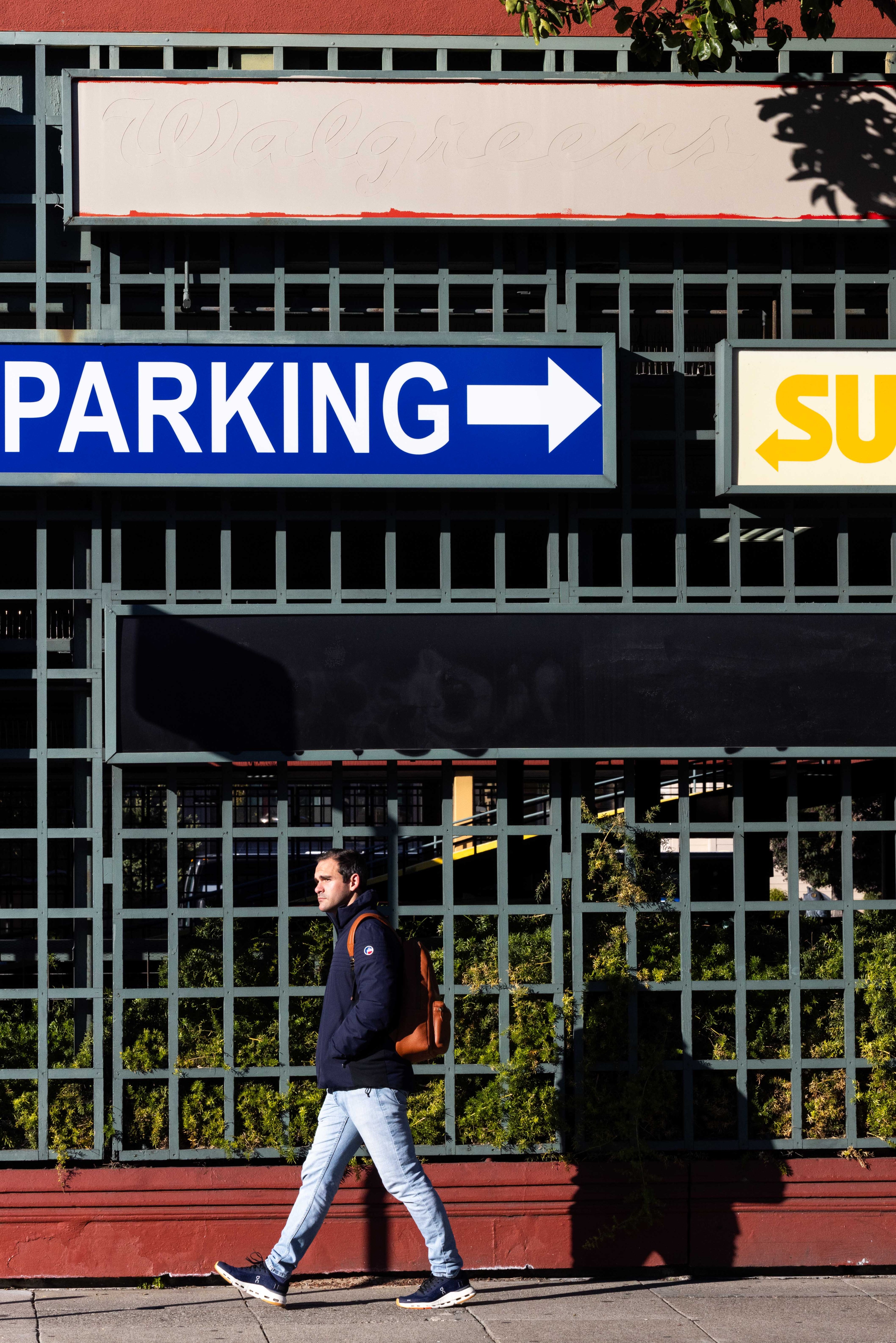 A man with a backpack walks past a grid-patterned structure. Above him, a blue sign says &quot;Parking&quot; with a right arrow, and a partial yellow sign points left.