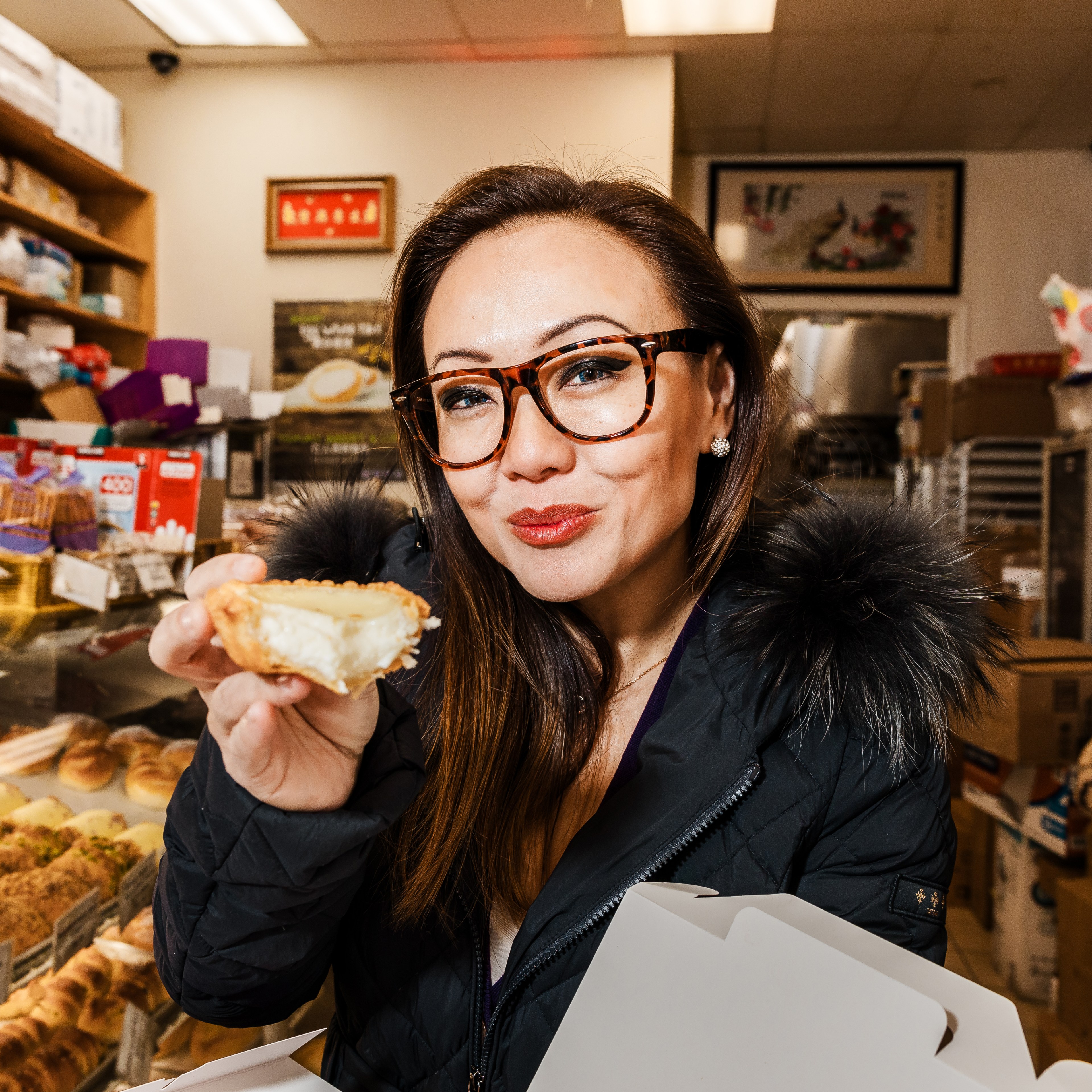 A person in a warm coat and glasses is inside a bakery, smiling while holding a partially eaten pastry. The background shows shelves filled with baked goods.