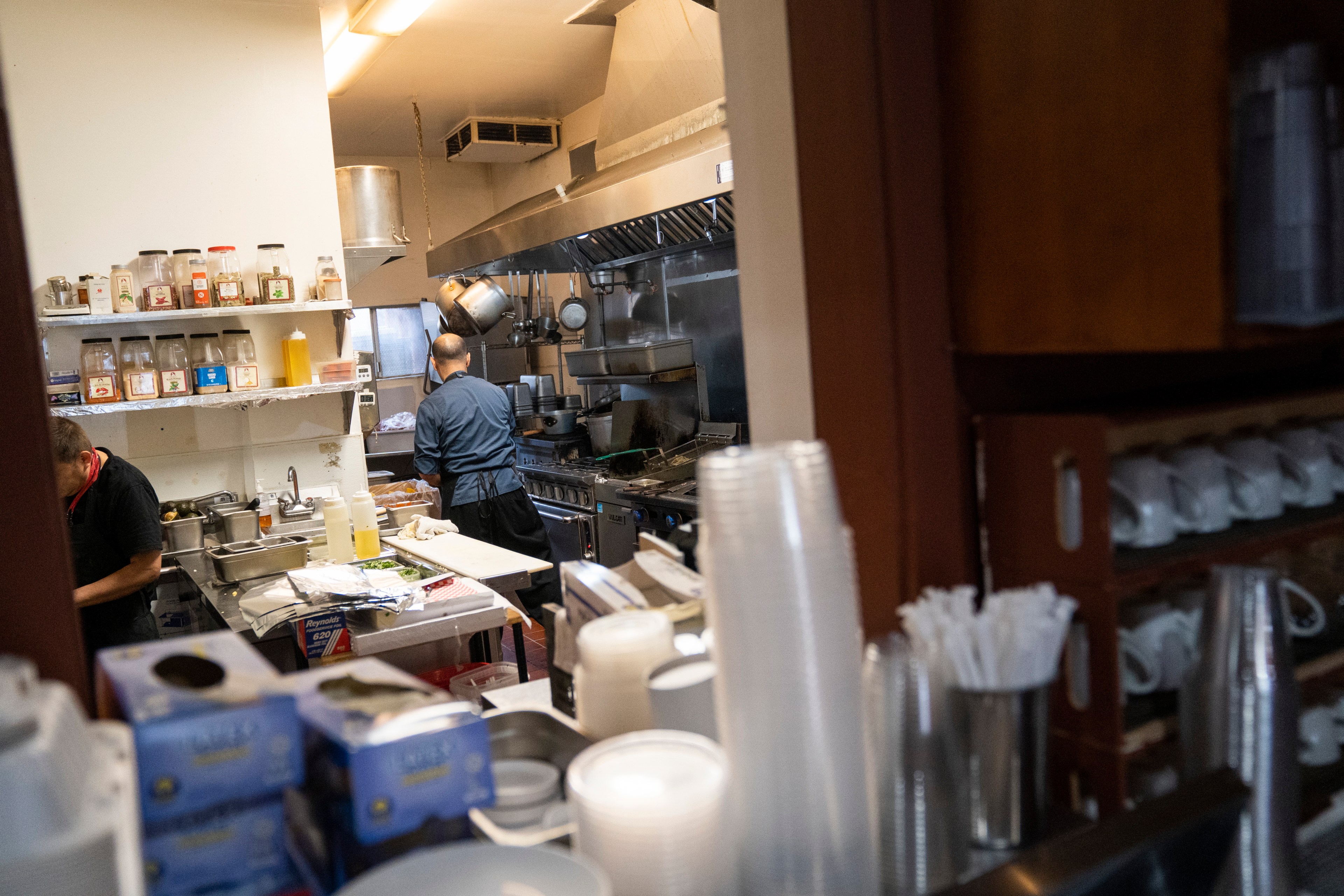 A busy restaurant kitchen with two people preparing food, shelves of spices, cooking utensils, and stacked disposable cups in the foreground.