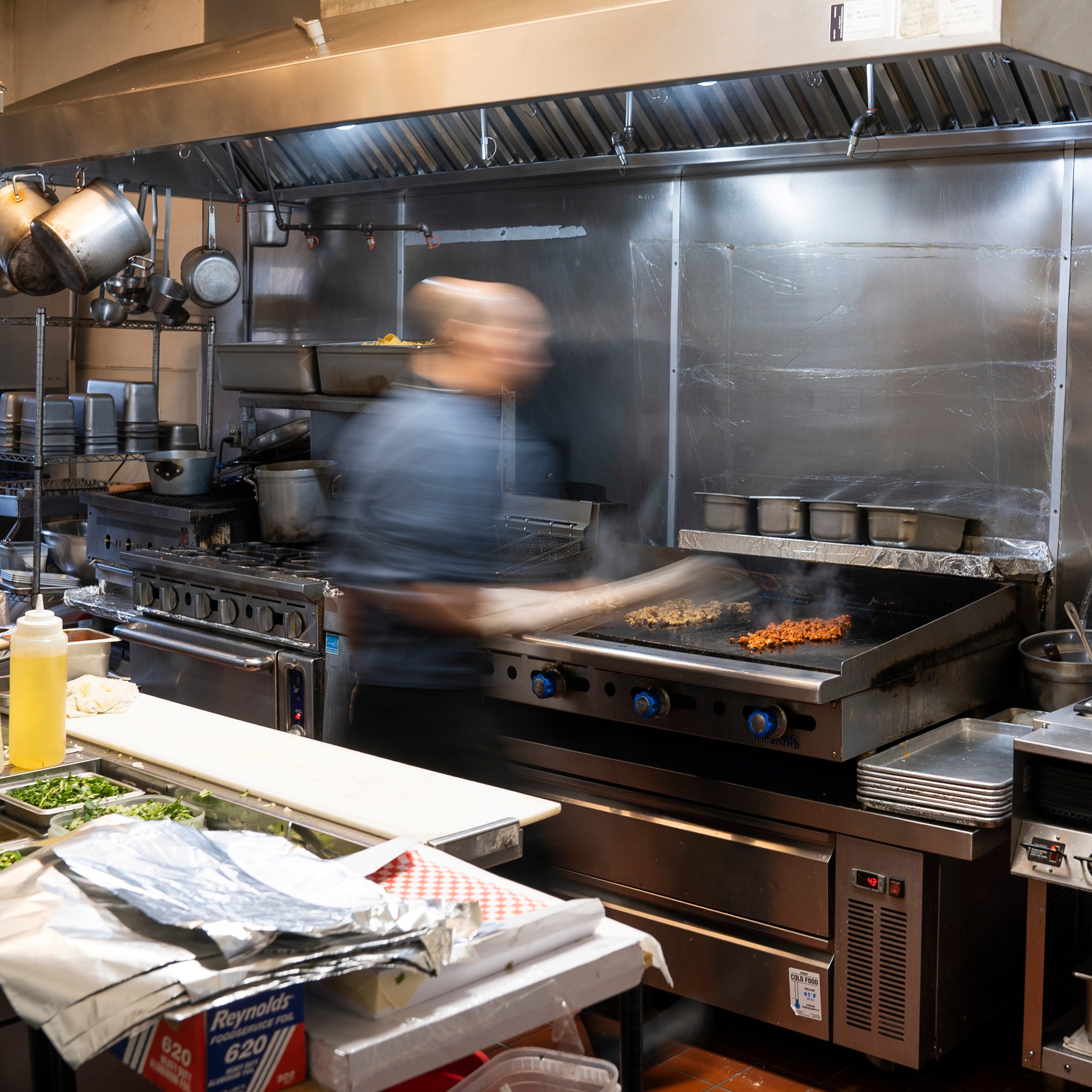 A chef in a bustling kitchen is cooking on a large grill, with lots of metal cookware around. Ingredients are prepared on the counter, and steam rises from the food.