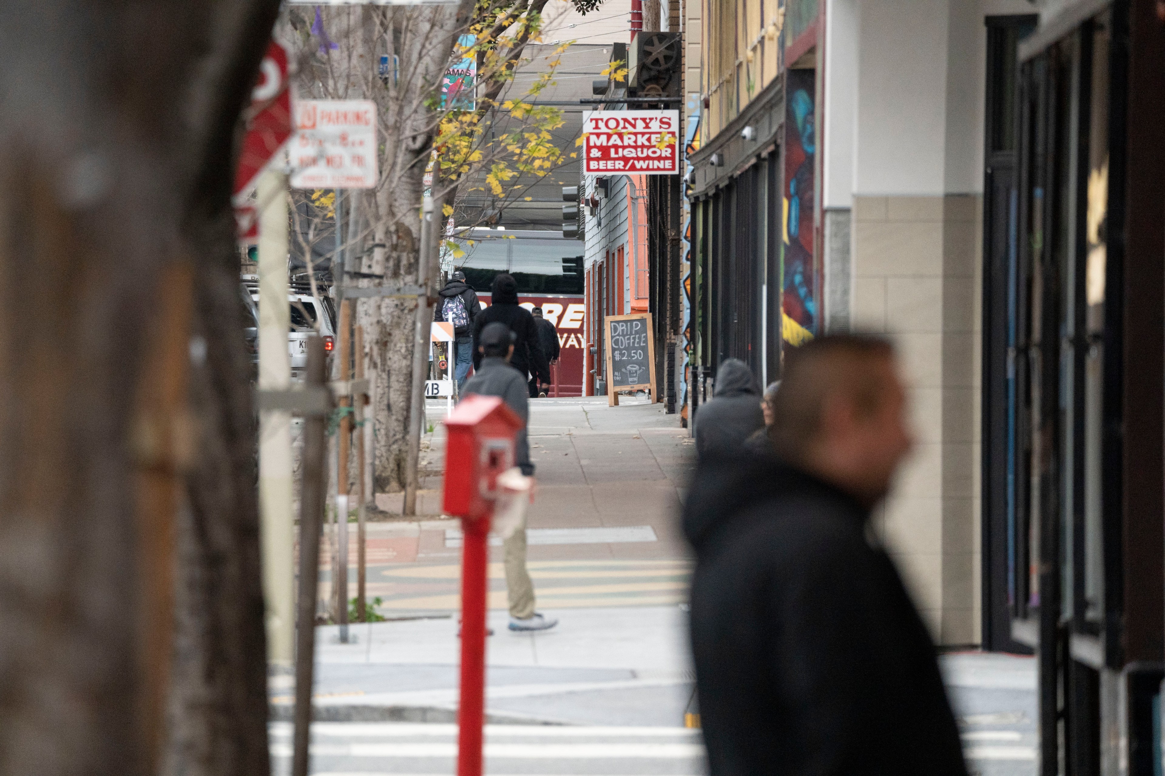 A bustling city sidewalk is lined with trees, people in jackets walk past shops. A &quot;Tony's Market &amp; Liquor&quot; sign is visible, with a board for drip coffee.