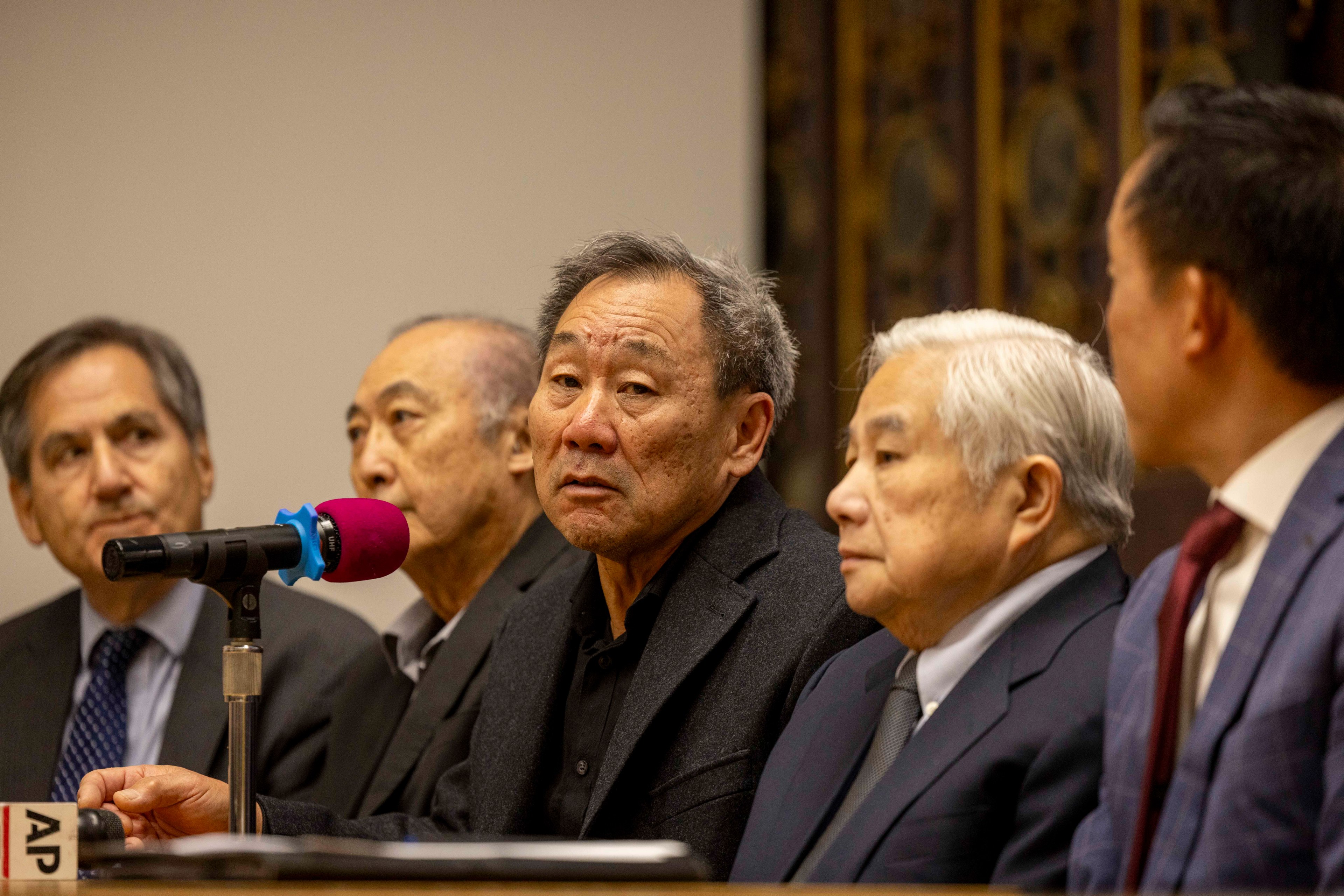 Five men in suits sit at a panel with a microphone on the table. They appear serious and are focused on the speaker who is using the microphone.