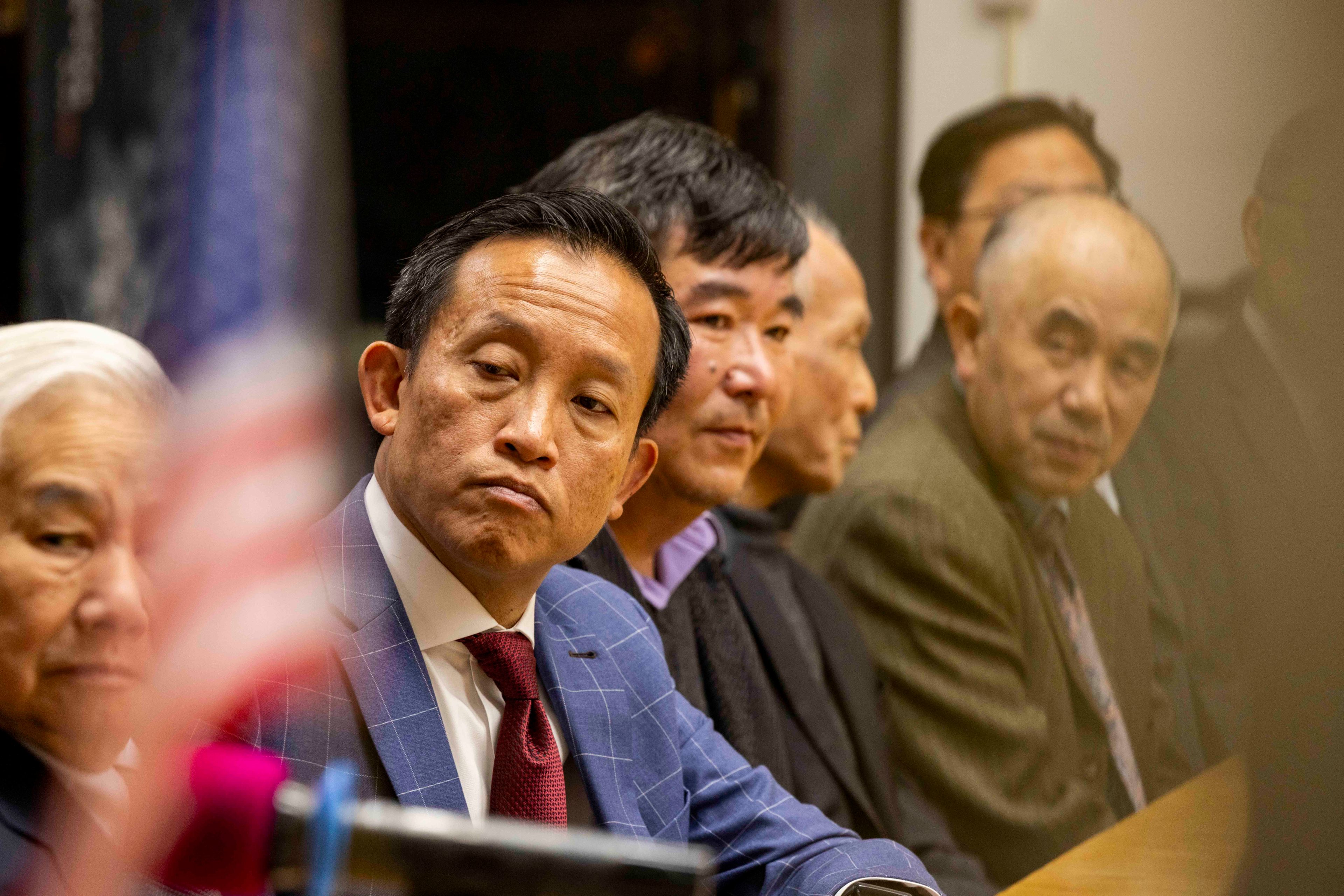 A group of men in suits sit at a table in a meeting room. The focus is on one man in a blue suit with a red tie, appearing attentive, with a blurred flag in the foreground.