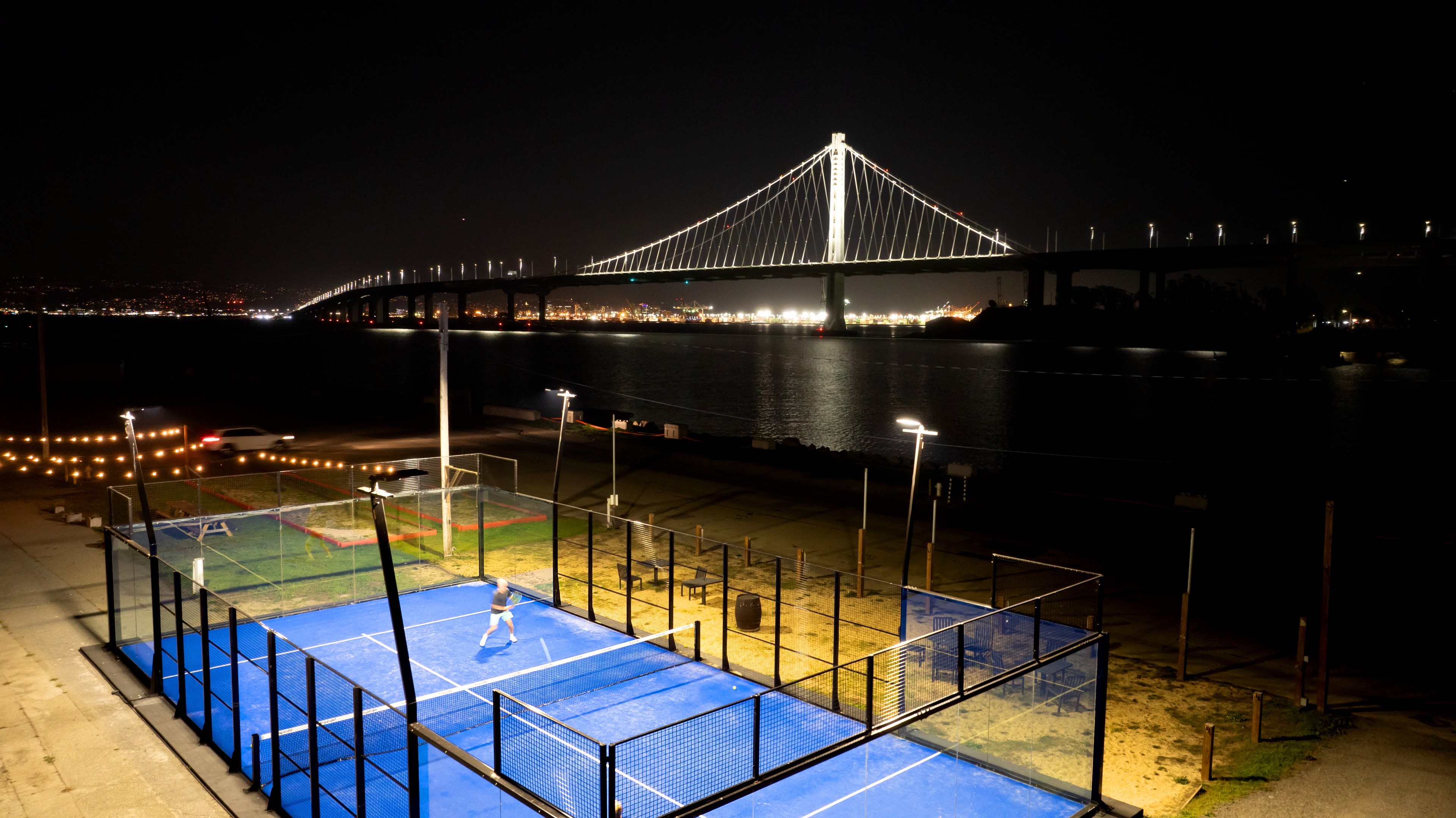 A person plays on a lit blue padel court at night. In the background, a brightly illuminated bridge spans across a body of water.