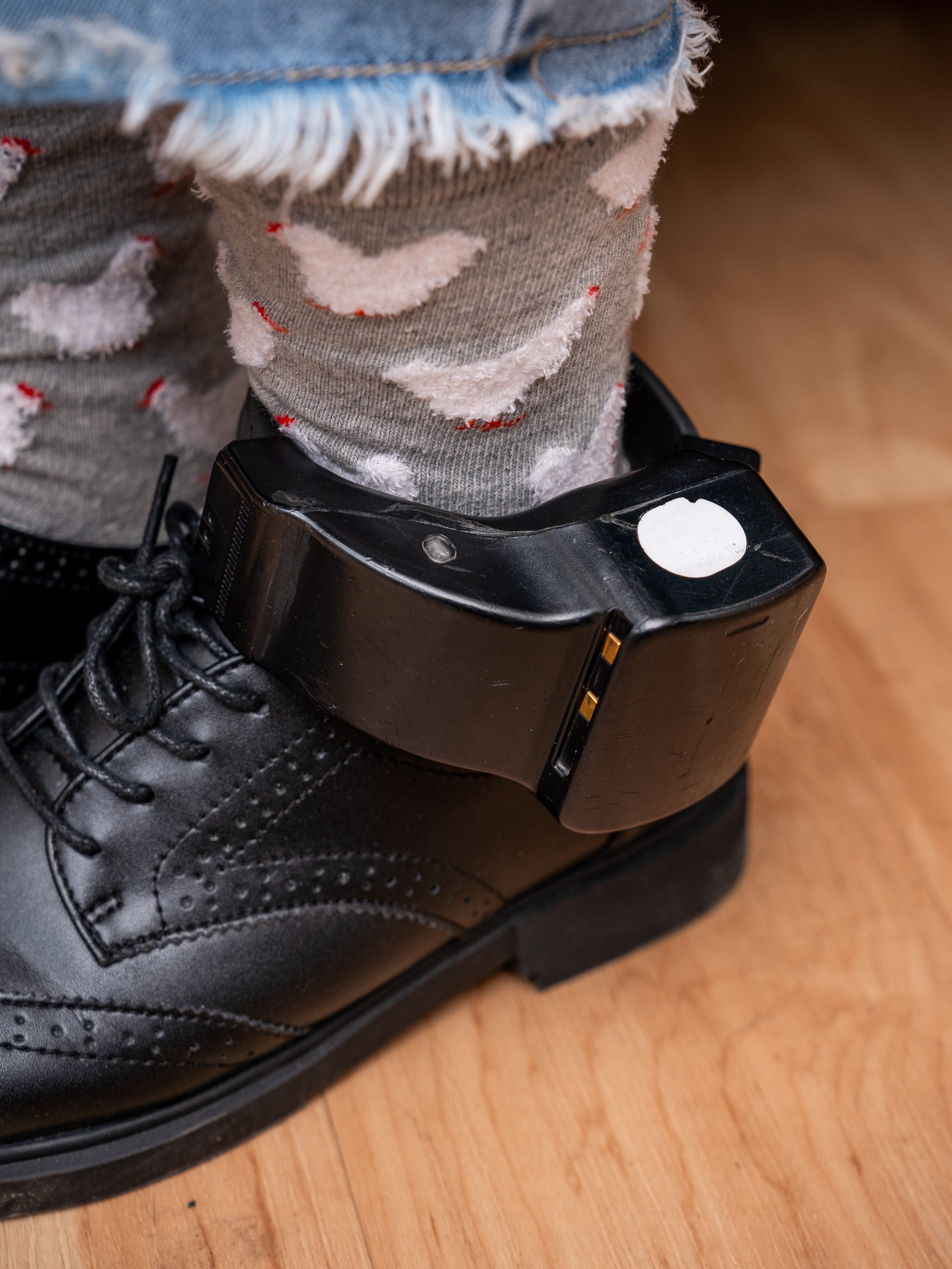 A person wears a black leather shoe with an ankle monitor. The visible sock is gray with white chickens, and the hem of frayed blue jeans is seen.