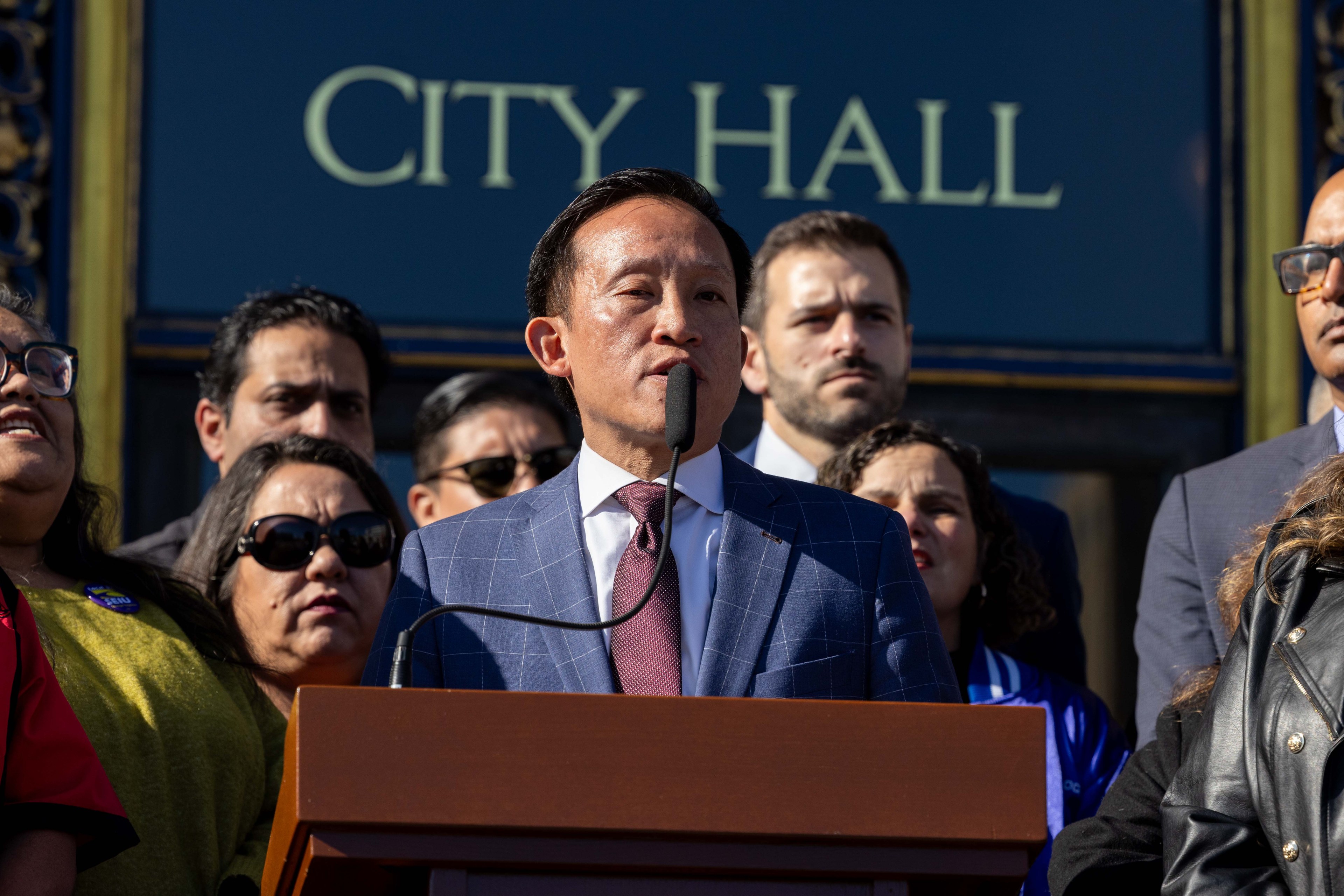 A person in a blue suit speaks at a podium outside a building marked "City Hall." Several people stand behind, listening attentively.