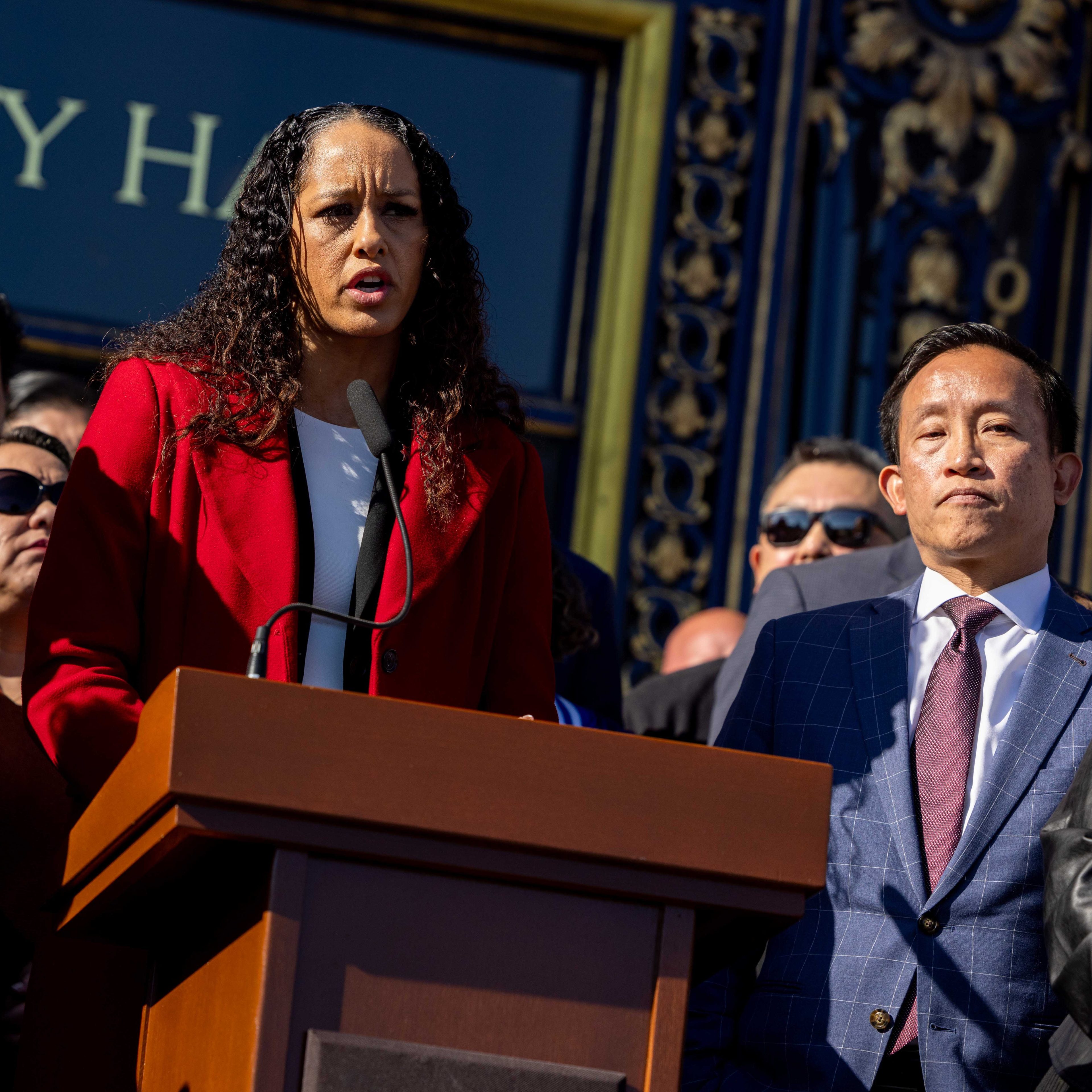 A woman in a red coat is speaking at a podium with a microphone, while a man in a suit stands beside her, both surrounded by a group of people.