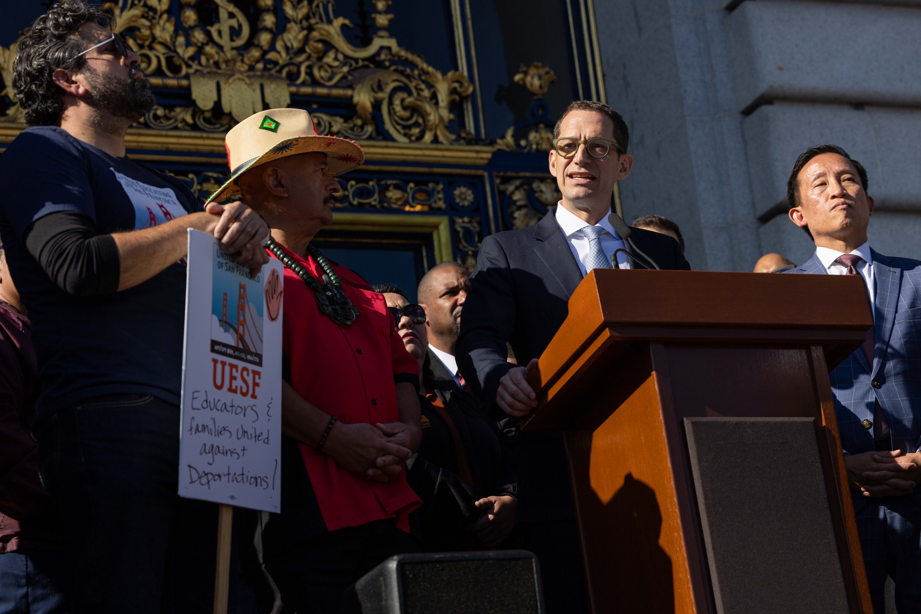 A man in a suit speaks at a podium, surrounded by others. One holds a sign reading &quot;UESF Educators &amp; families united against deportations&quot; with a Golden Gate Bridge image.