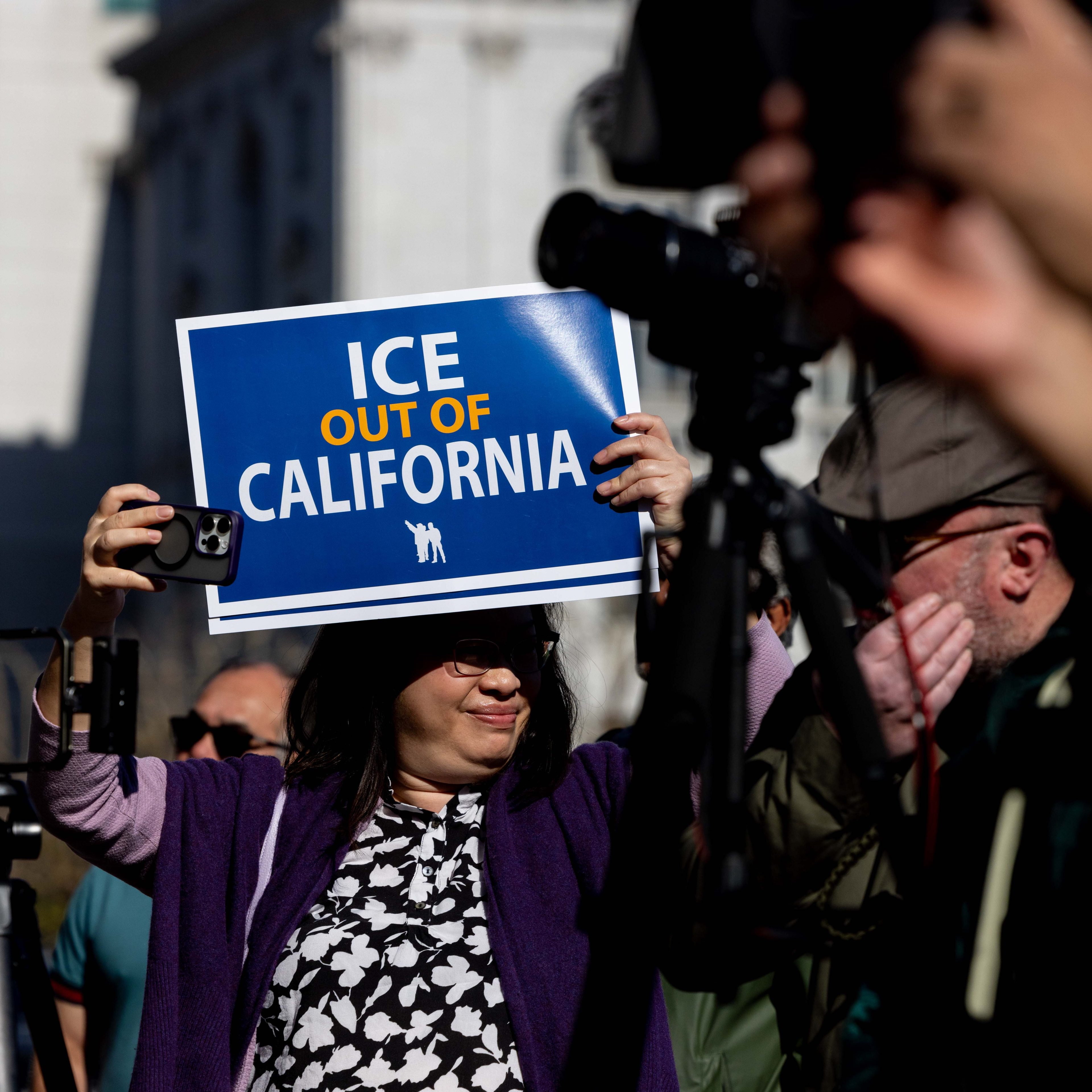 A person holds a blue sign reading &quot;ICE OUT OF CALIFORNIA&quot; at a protest. They wear glasses and purple clothing, while others around them also appear engaged.