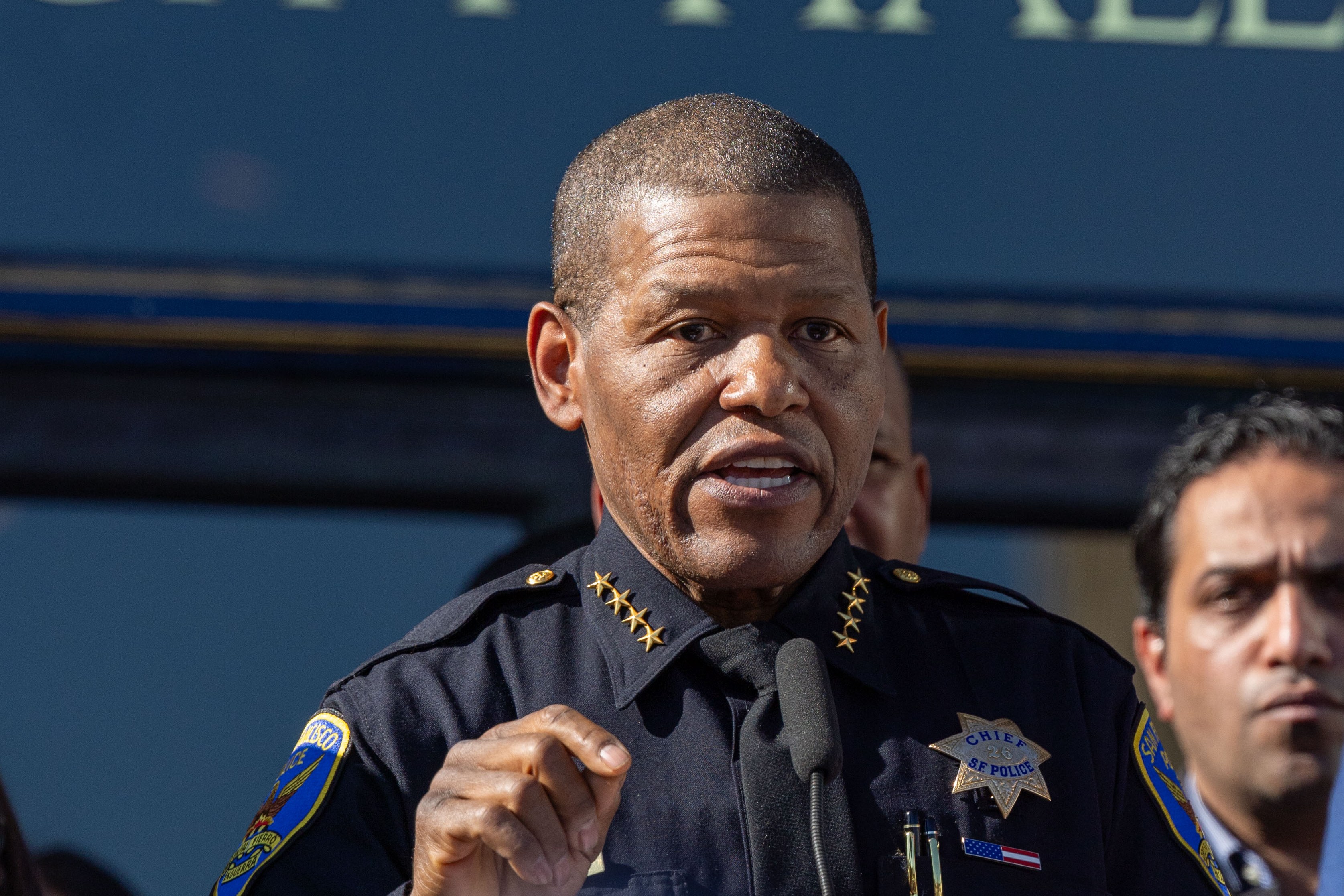 A person in a police uniform speaks at a podium outdoors, in front of a building labeled "City Hall," with other people standing behind them.