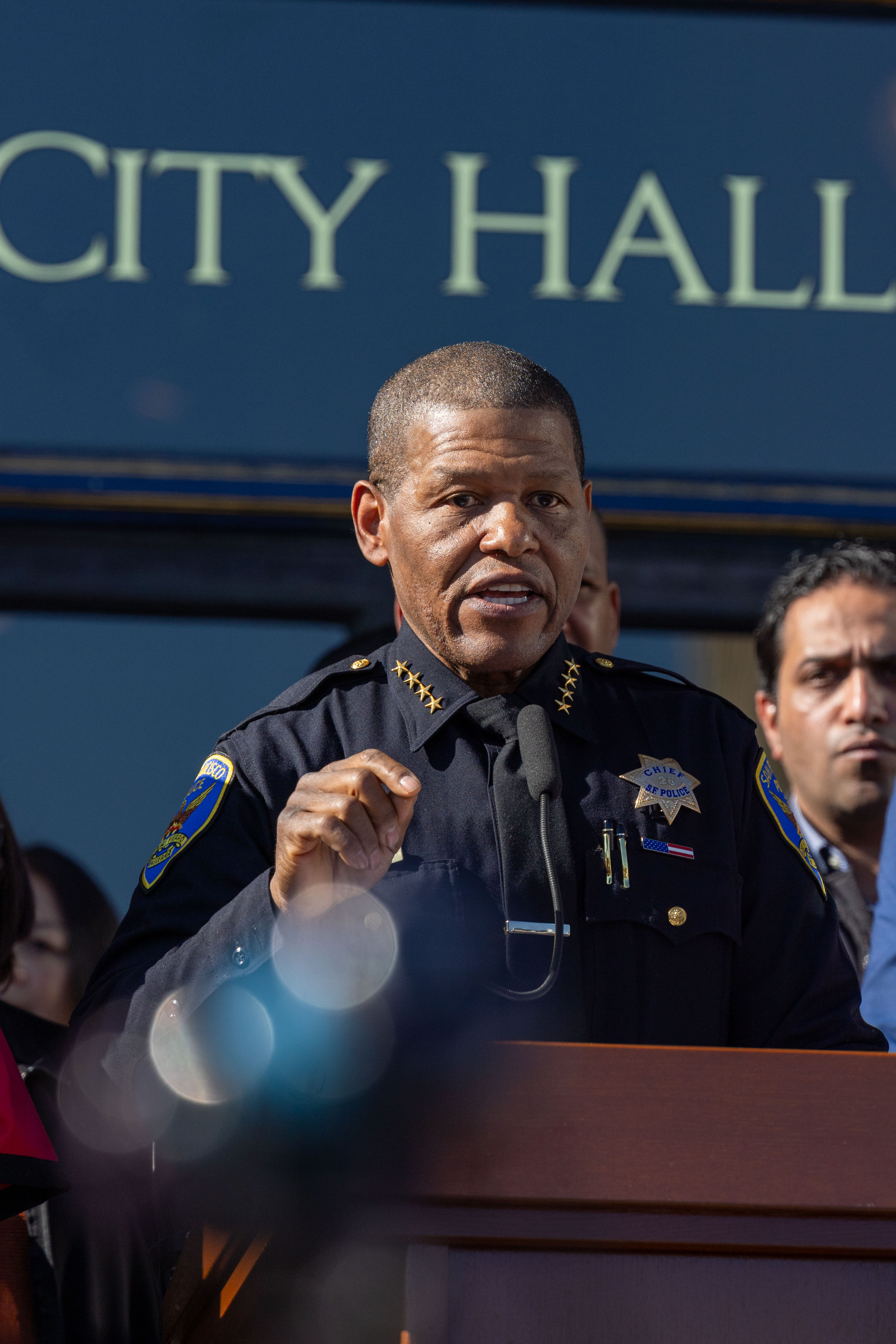 A person in a police uniform speaks at a podium outdoors, in front of a building labeled "City Hall," with other people standing behind them.