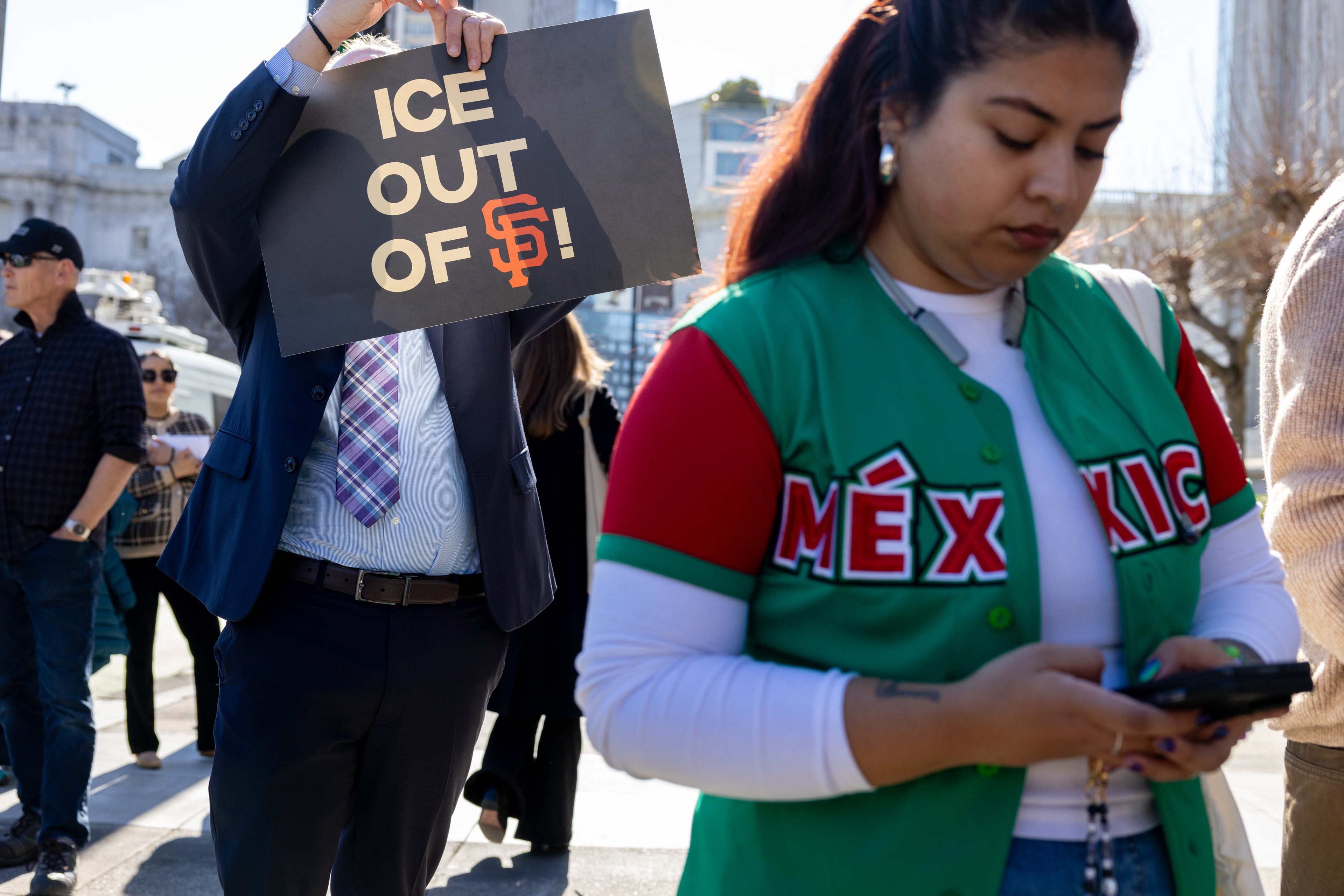 A person holds a sign reading "ICE OUT OF SF!" at a protest. Another individual nearby wears a green jersey with "MÉXICO" and is focused on a phone.