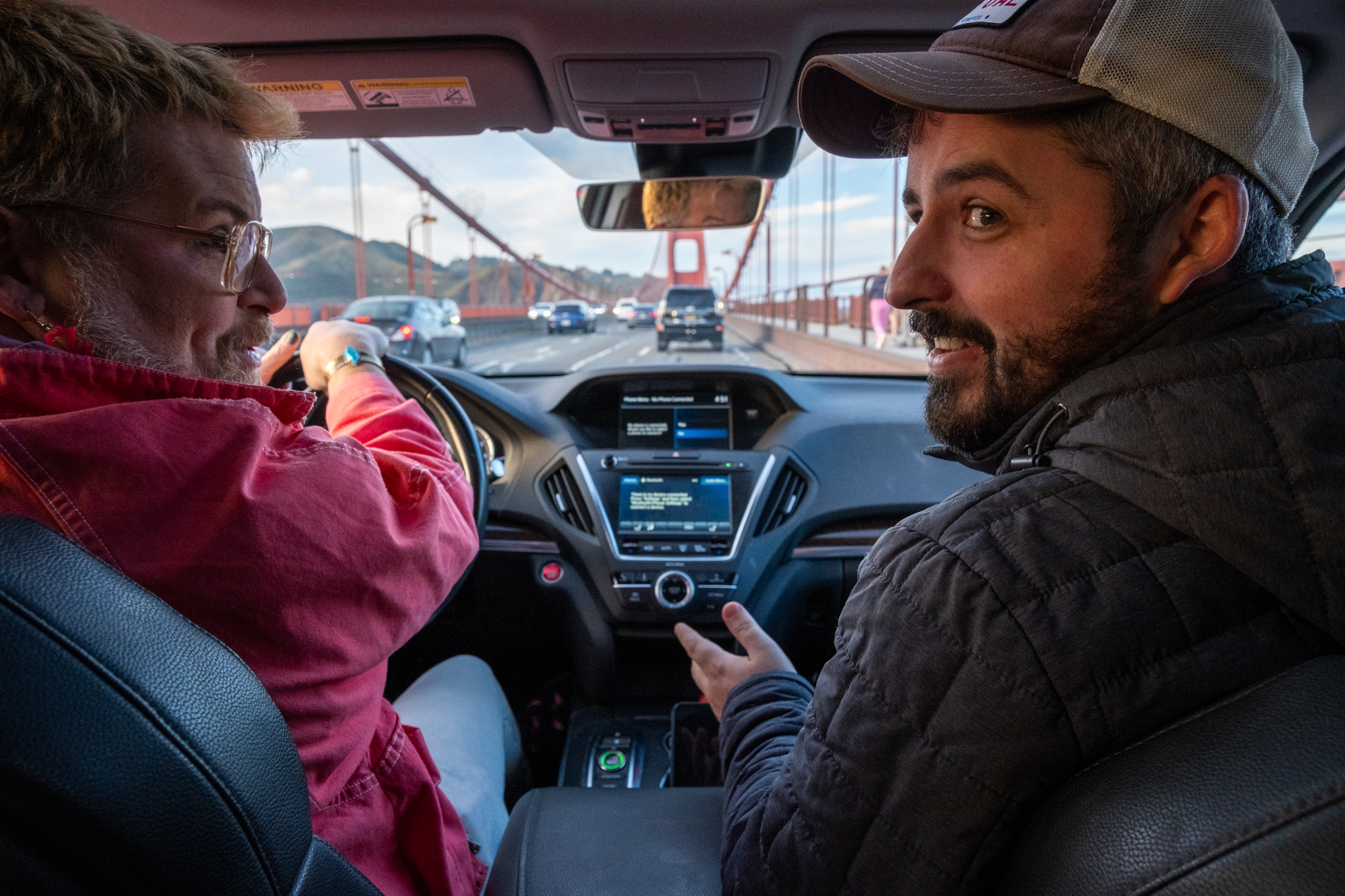 Two people sit in a car on a bridge, with one driving and the other smiling. The car's dashboard and the bridge structure are visible through the windshield.