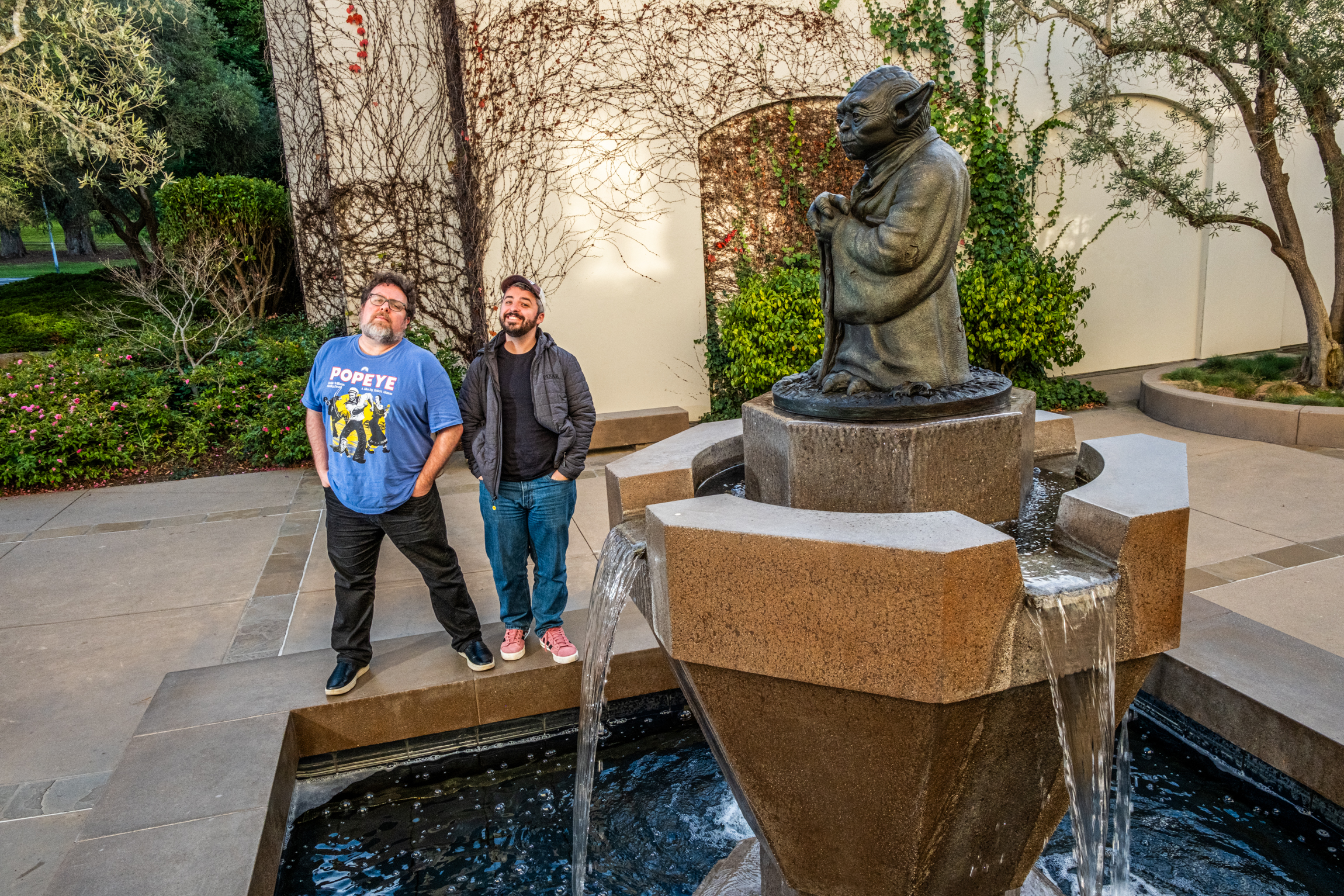 Two people stand near a fountain with a statue of a wise-looking creature. The area is outdoors, flanked by greenery and a wall adorned with vines.