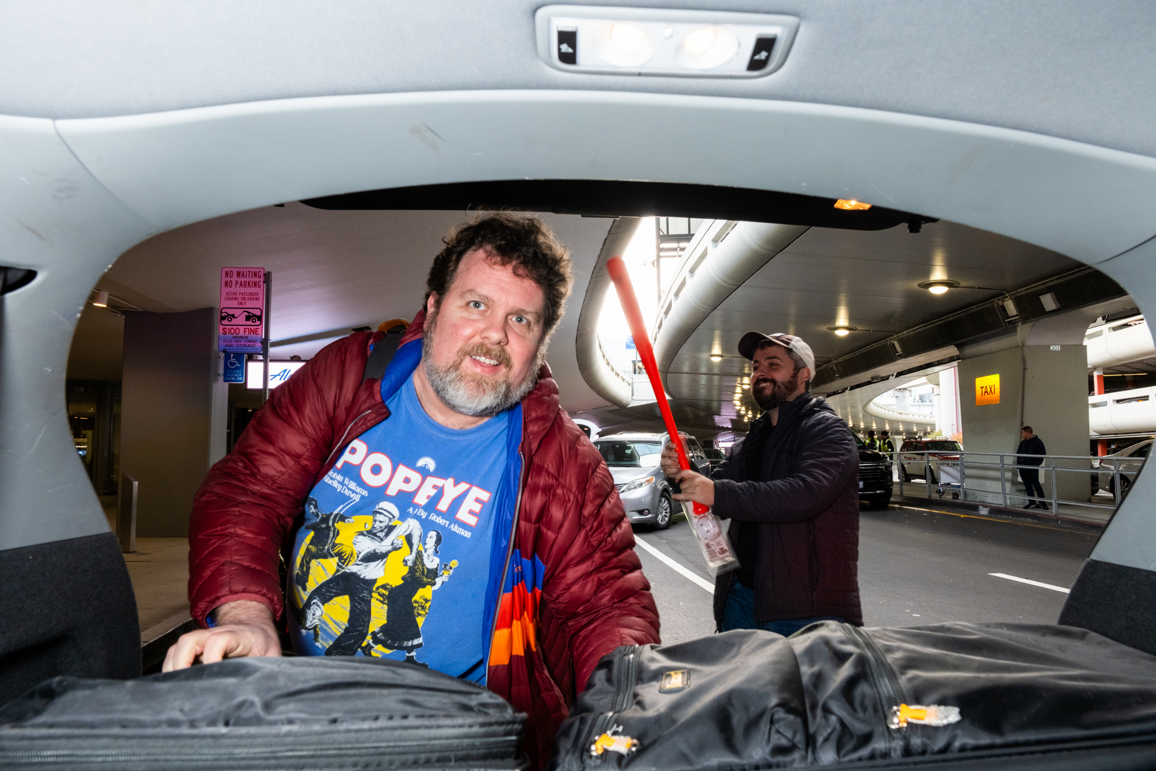 A man in a red jacket is leaning into a car trunk, wearing a blue Popeye shirt. Another man holds a large red bat behind him, and they are on a busy street.