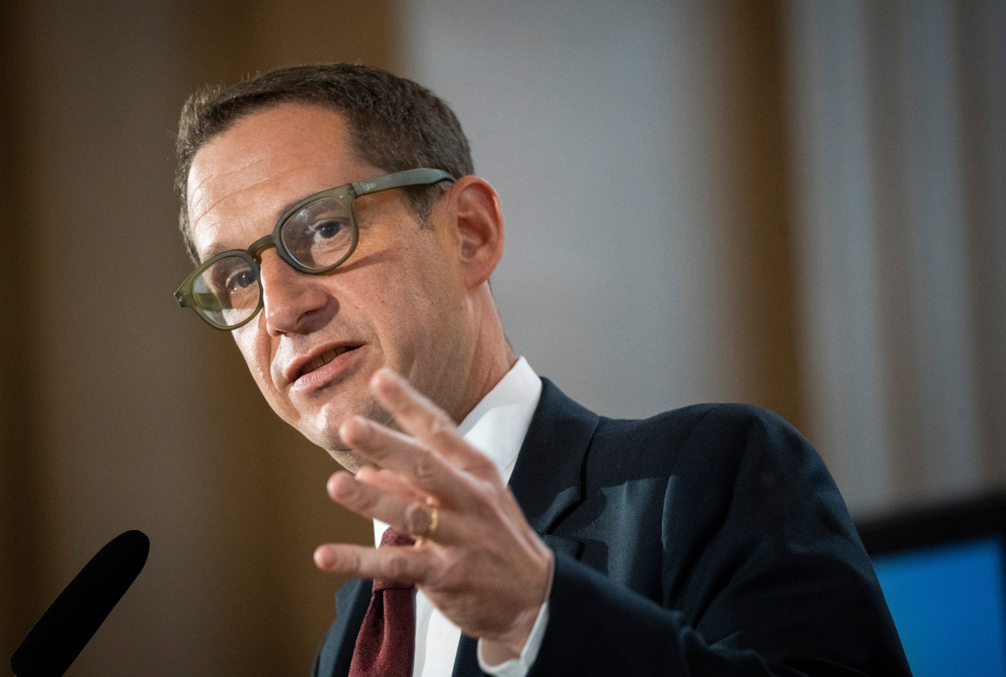 A person with glasses is speaking, gesturing with their hand. They wear a suit and tie, and there is a microphone visible. The background is blurred.