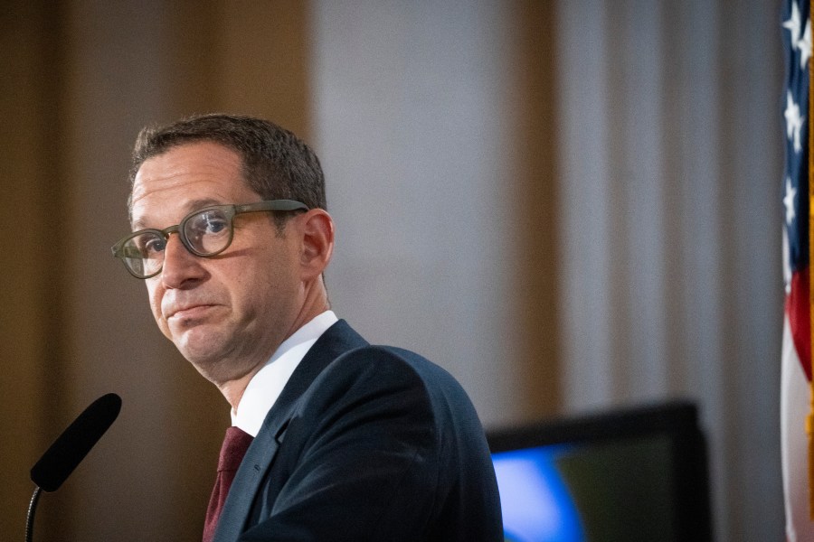 A man in glasses and a suit stands at a microphone, with an American flag partially visible to the right. His expression is serious, and the background is blurred.