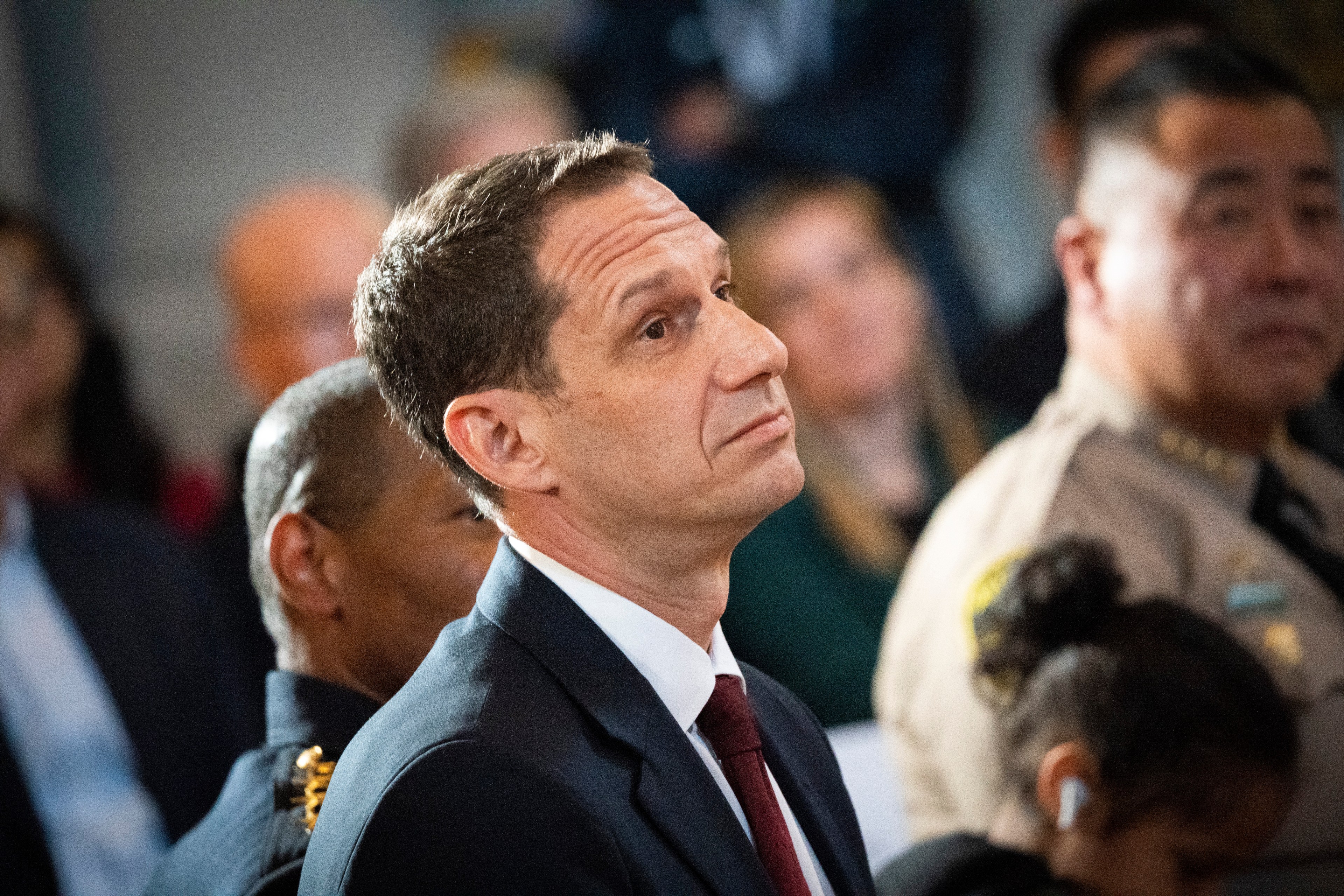 A man in a suit and tie looks upward, surrounded by people in formal attire, including a uniformed officer and a person in a tan suit. The setting appears formal.