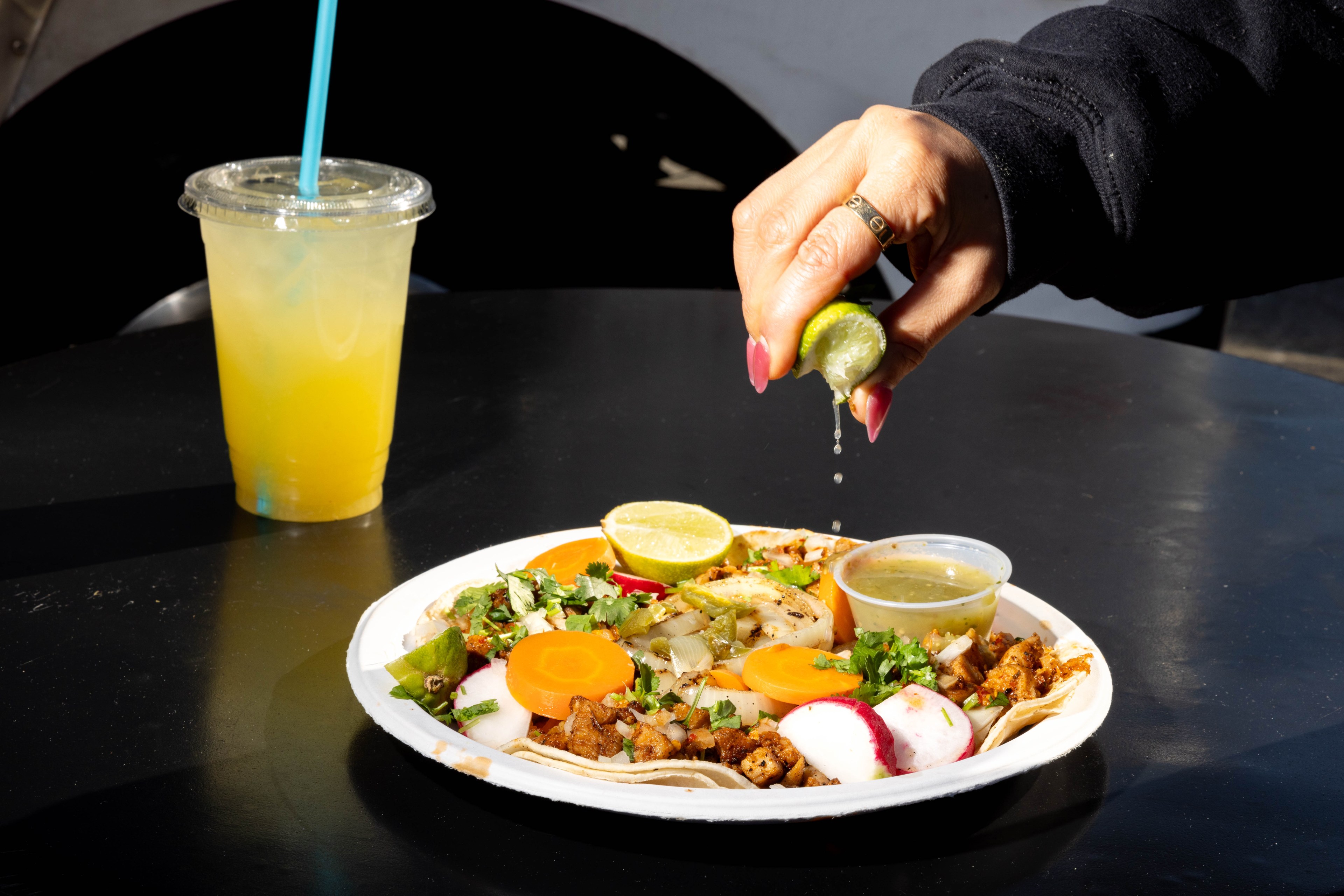 A hand squeezes lime over tacos with radish, carrot, and cilantro on a plate; a cup of yellow drink with a blue straw is beside the plate.