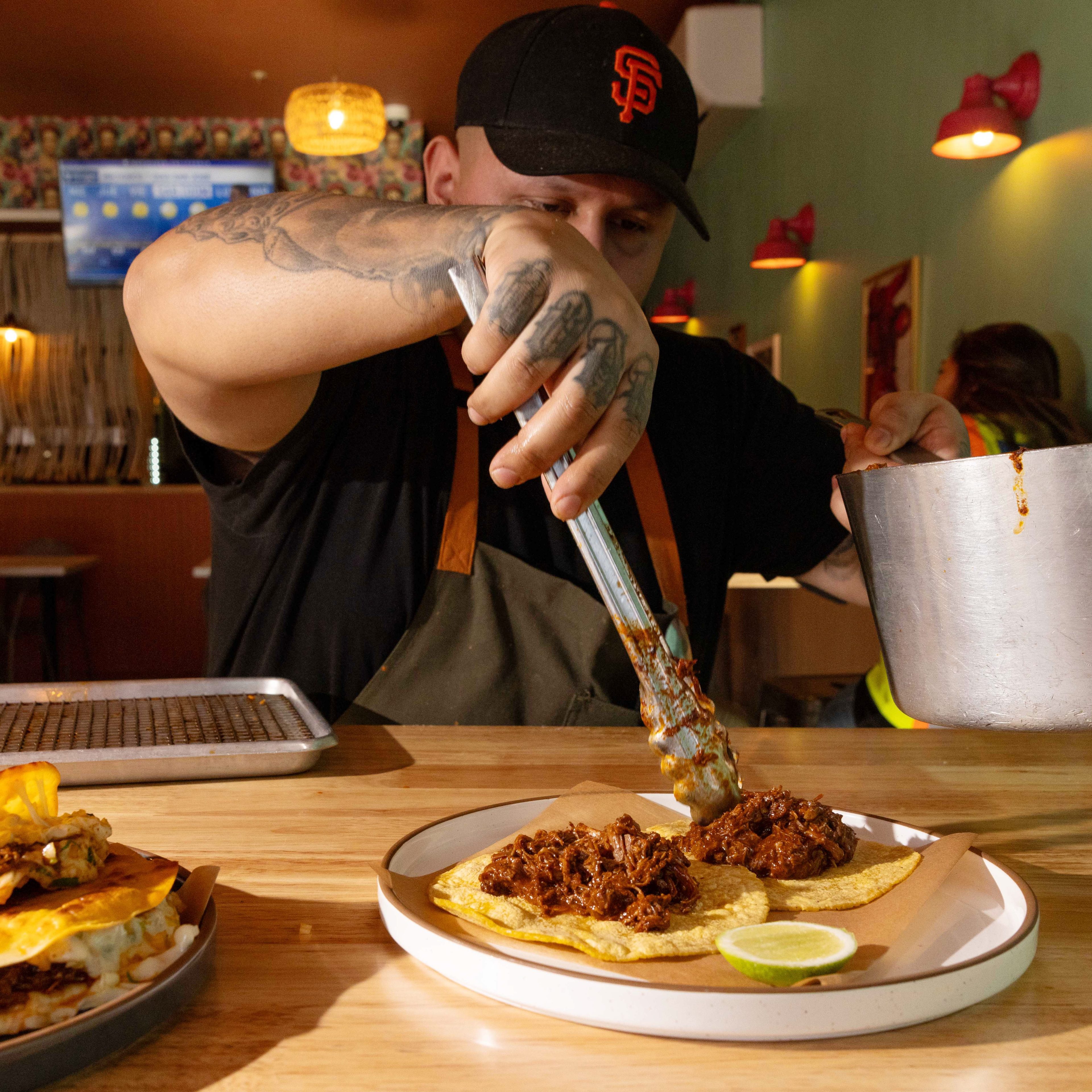 A person wearing a black cap and apron uses tongs to place seasoned meat onto tortillas on a plate, with a lime slice nearby on the table.