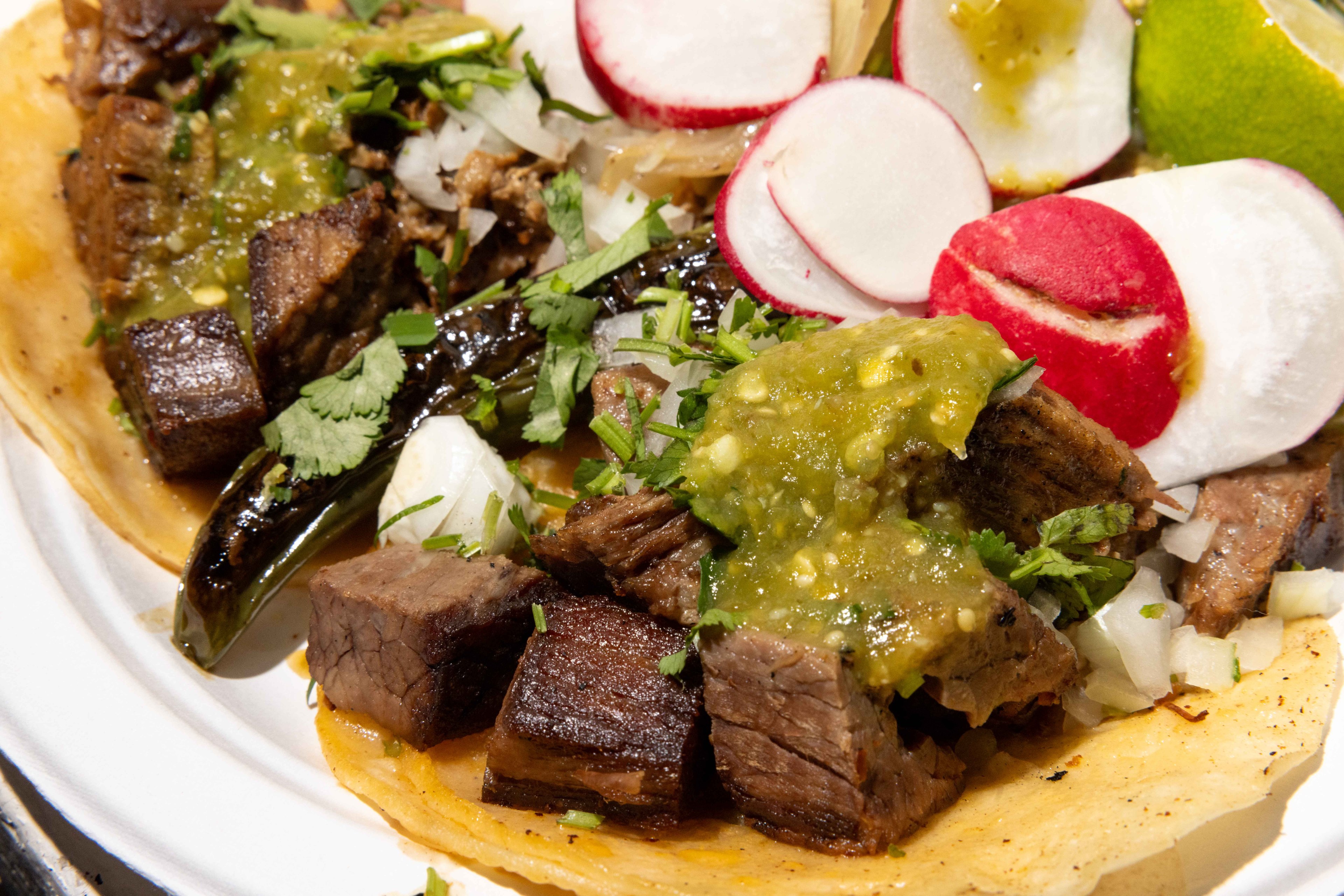 This image shows a taco topped with grilled beef, green salsa, chopped onions, cilantro, sliced radishes, and a lime wedge on the side.