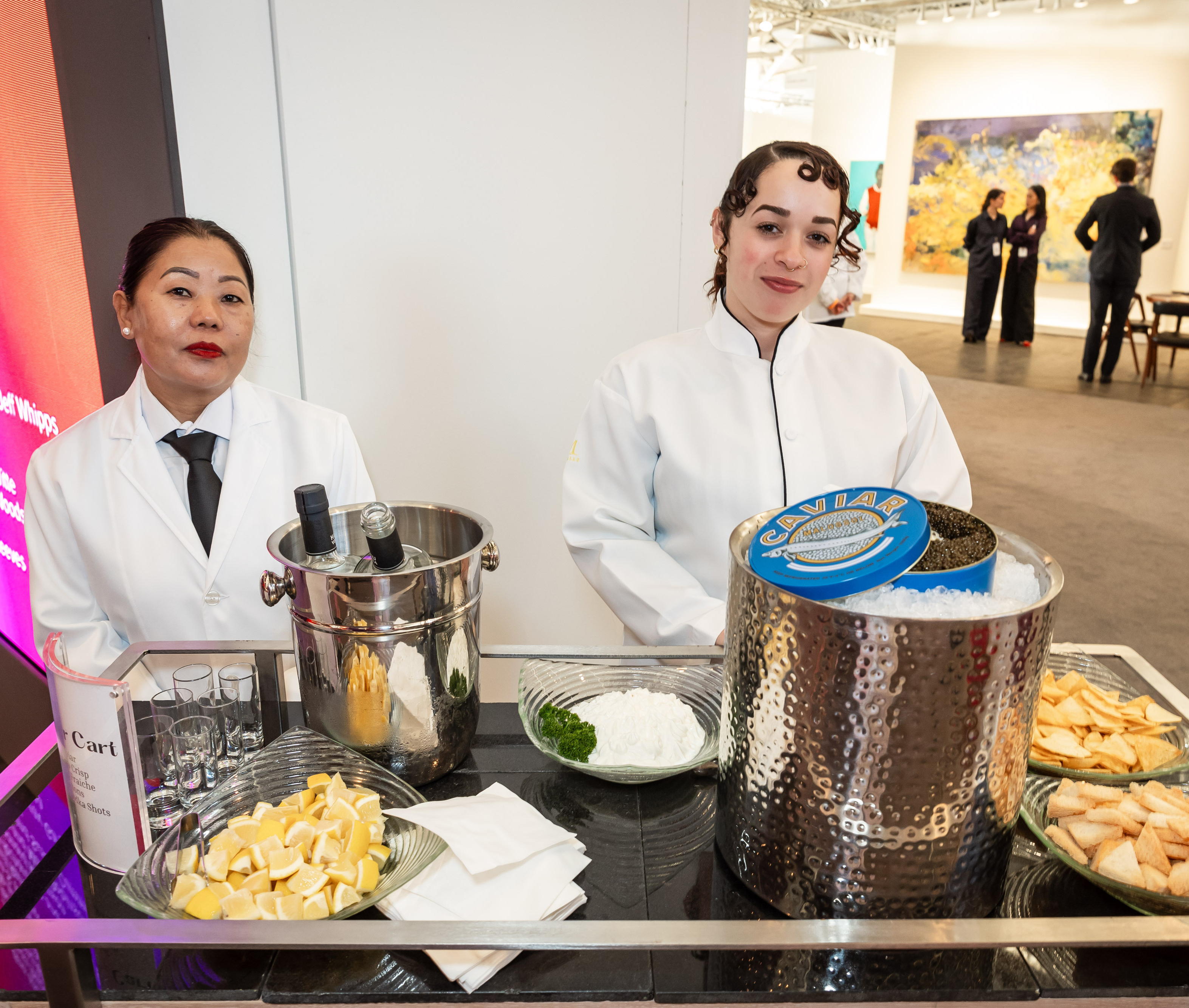 Two women in white uniforms stand behind a caviar cart with ice, lemons, parsley, and crackers. In the background is an art gallery setting.