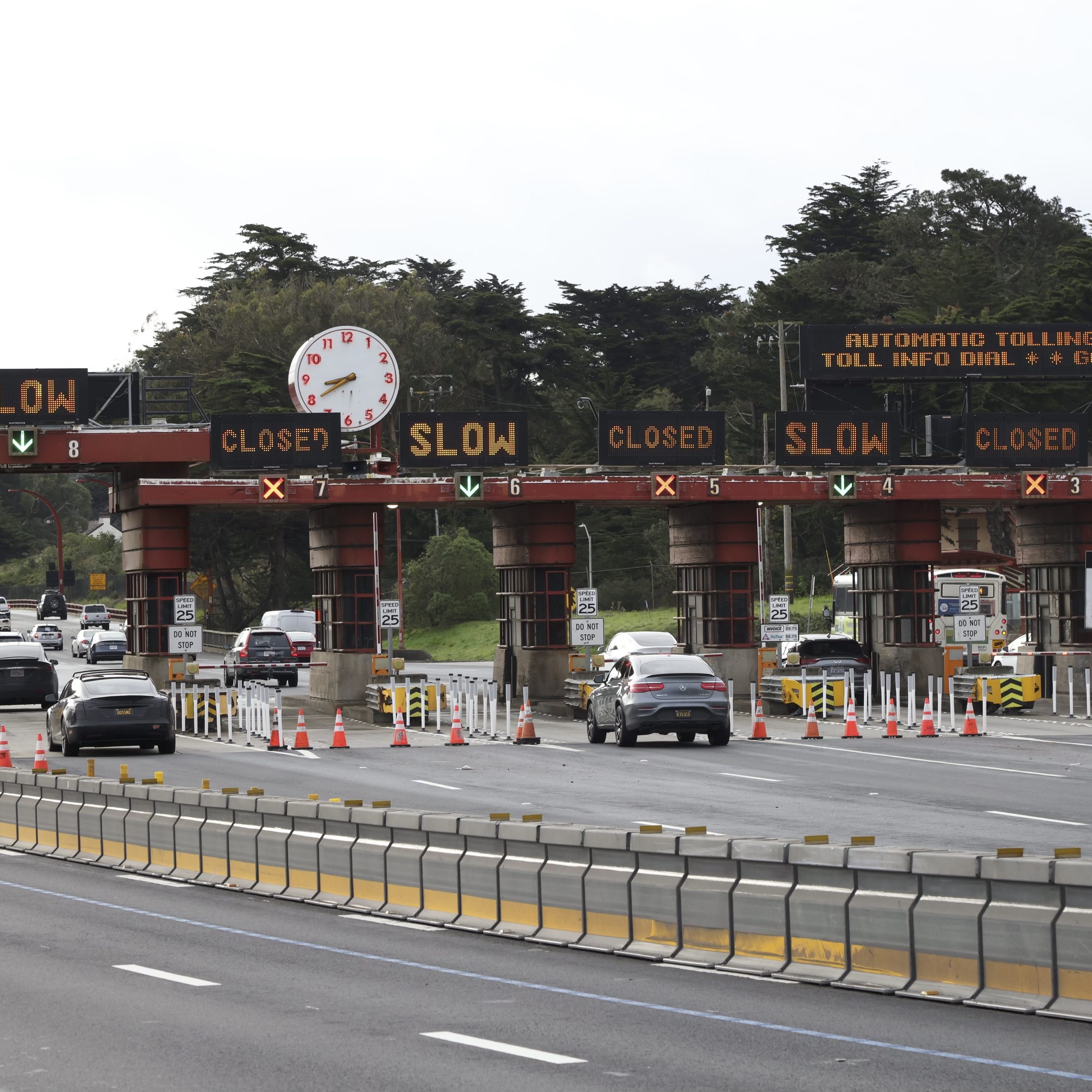 Cars are approaching a toll booth with signs saying &quot;SLOW&quot; and &quot;CLOSED.&quot; A large clock is displayed above the booth, surrounded by trees.