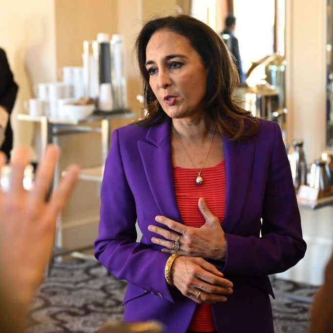 A woman in a purple blazer and red shirt is speaking to someone off-camera. She has a thoughtful expression and gestures with her hands. Others are in the background.