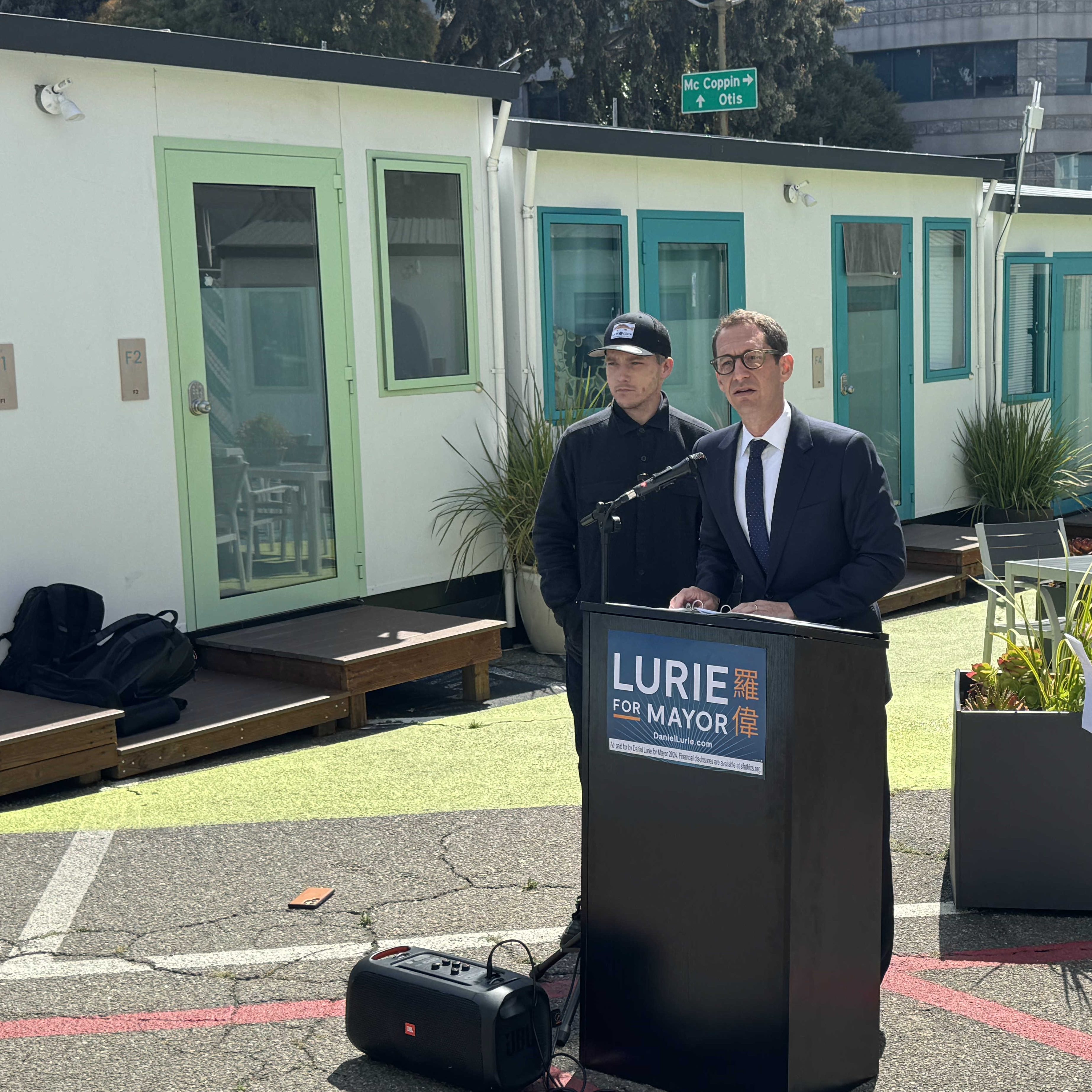 A man in a suit speaks at a podium labeled &quot;Lurie for Mayor,&quot; with two people nearby. Modular buildings and outdoor seating are visible in the background.