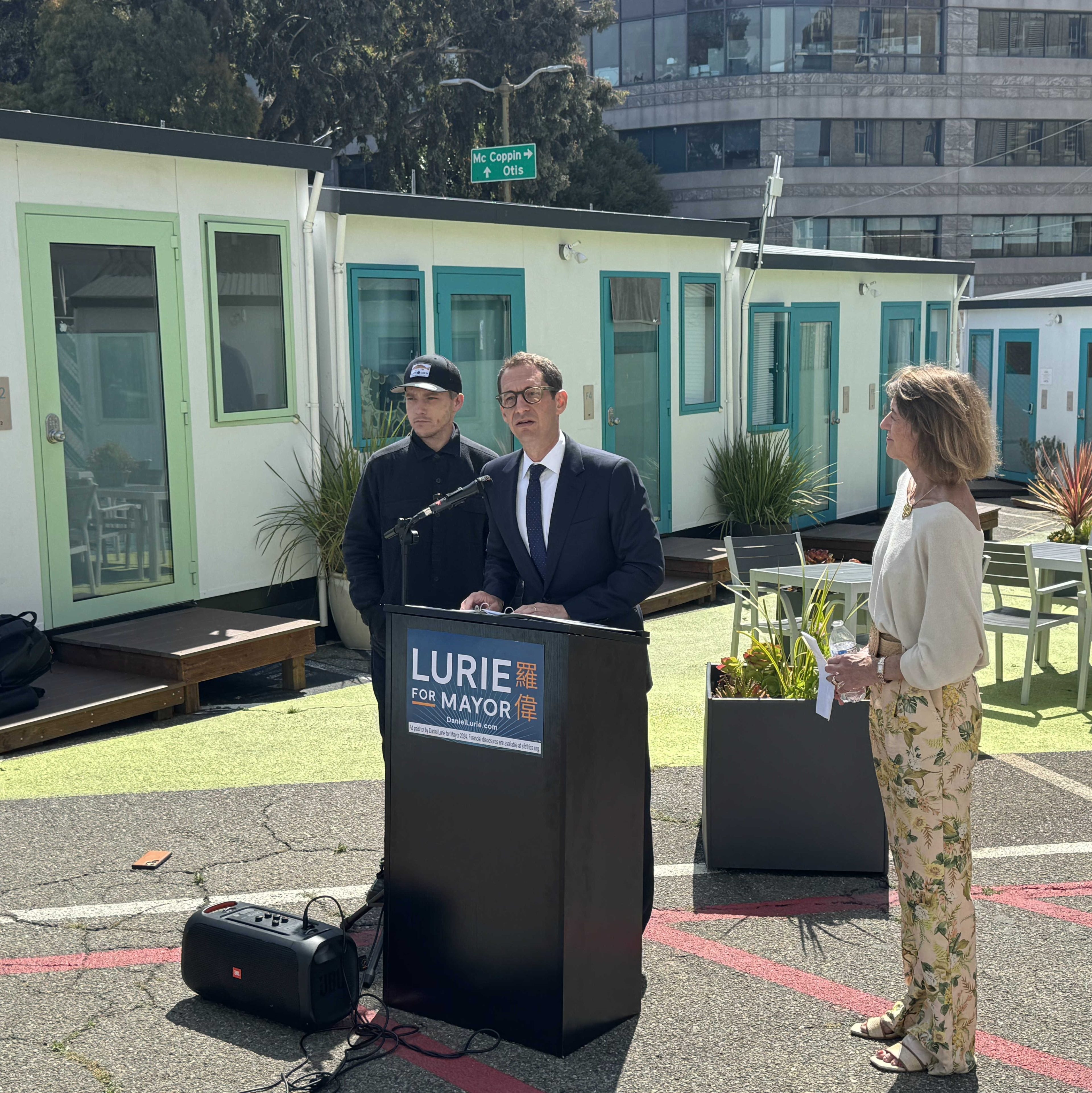 Three people stand outside small buildings, with one speaking at a lectern labeled &quot;Lurie for Mayor.&quot; The setting is urban, with plants and casual seating.
