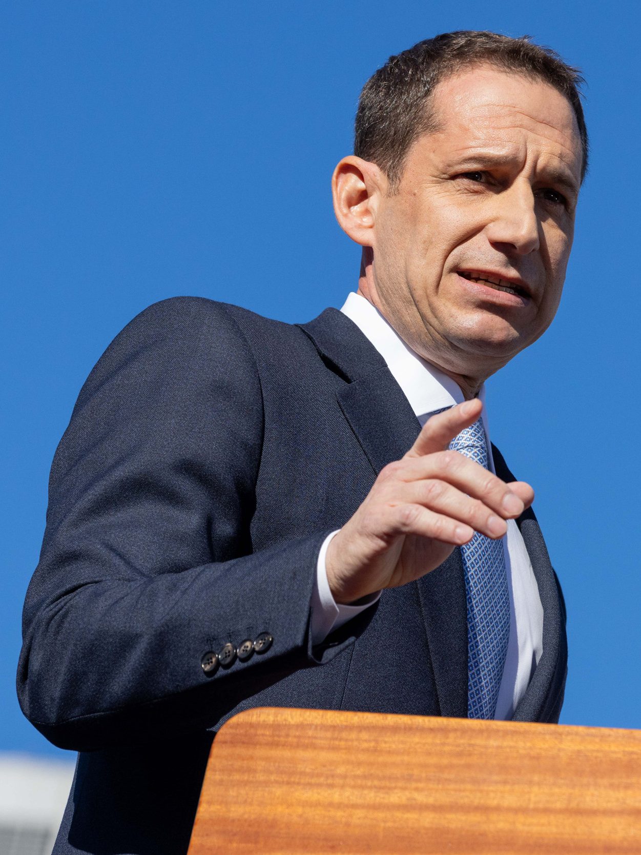 A man in a suit speaks at a podium with a microphone against a clear blue sky, gesturing with his hand.