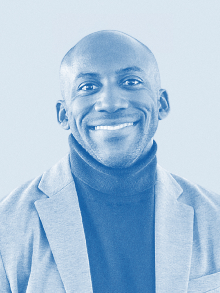 A person is smiling, wearing a turtleneck and blazer. The photo has a blue tint. The person is bald and appears to be facing the camera directly.