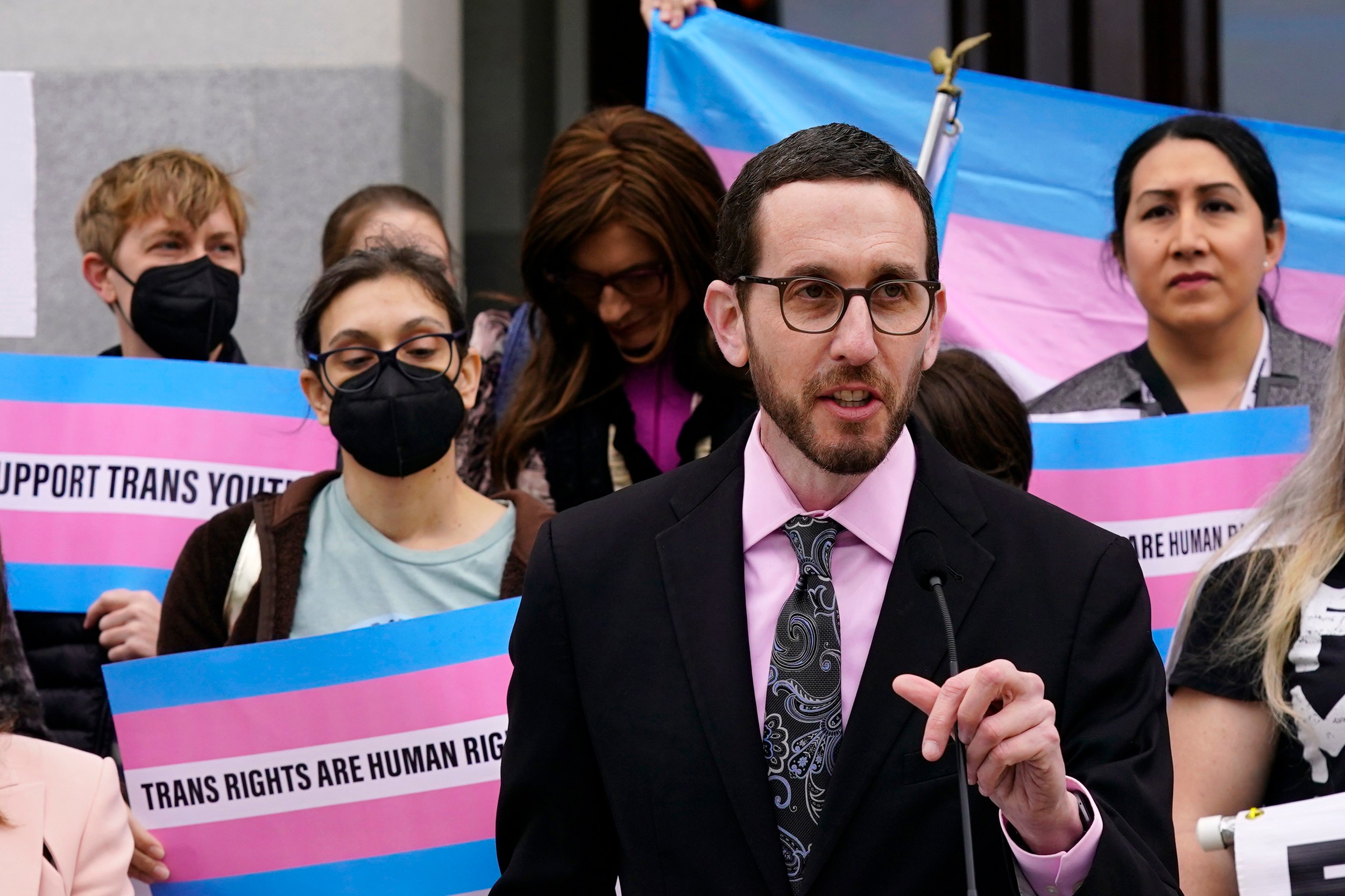 A person in a suit speaks at a podium with a crowd behind them holding pink and blue signs reading "Trans Rights Are Human Rights."