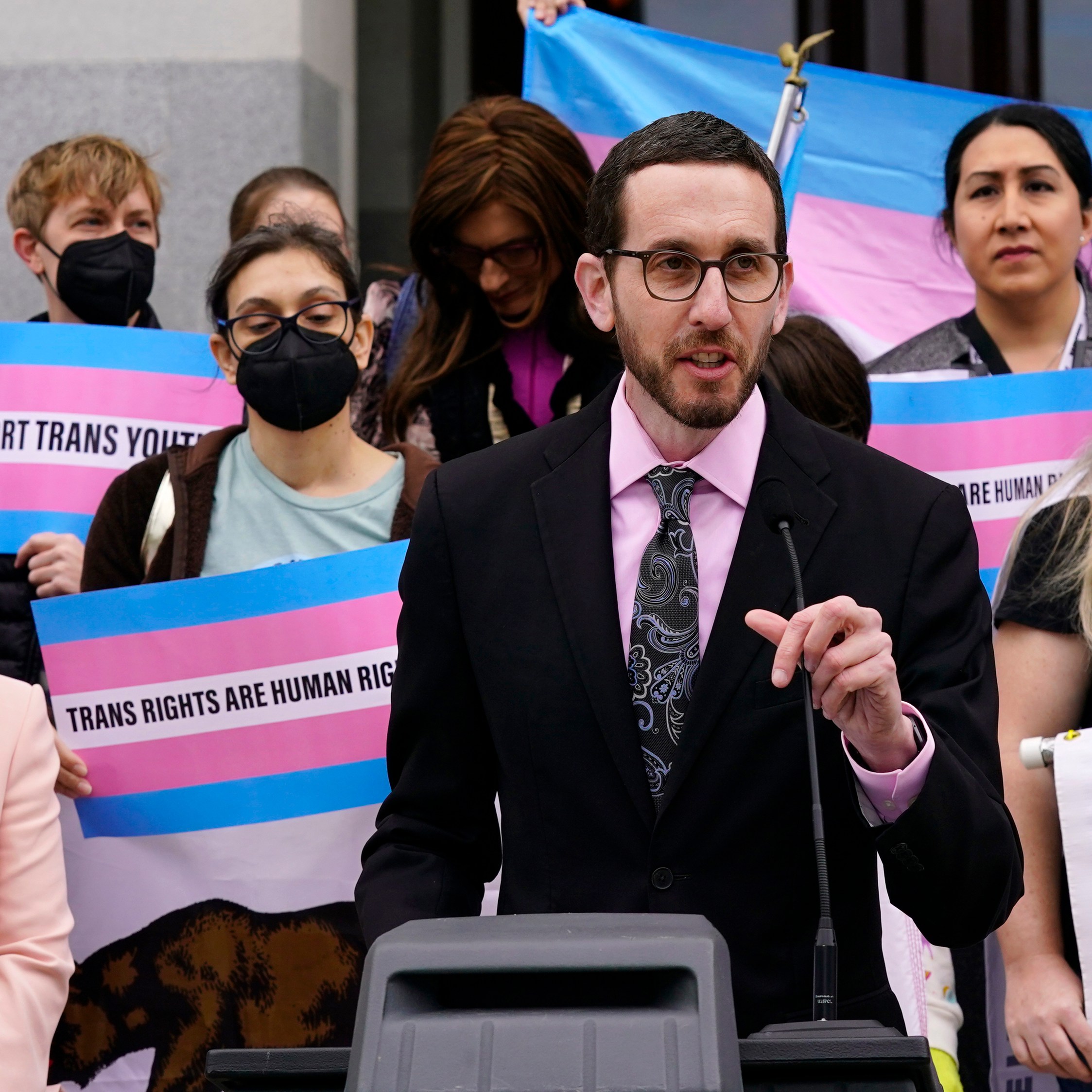 A person in a suit speaks at a podium with a crowd behind them holding pink and blue signs reading "Trans Rights Are Human Rights."