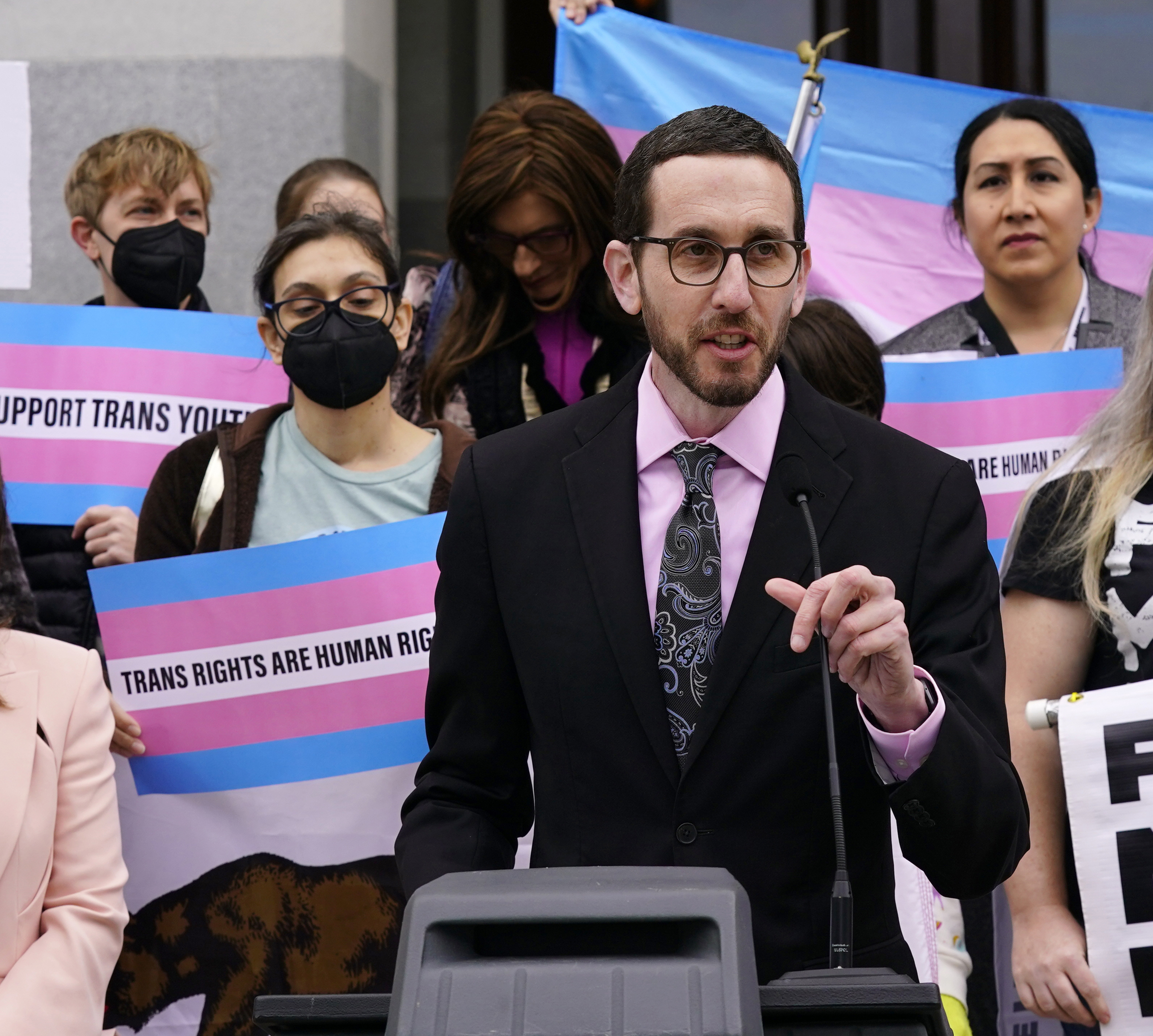 A person in a suit speaks at a podium with a crowd behind them holding pink and blue signs reading "Trans Rights Are Human Rights."