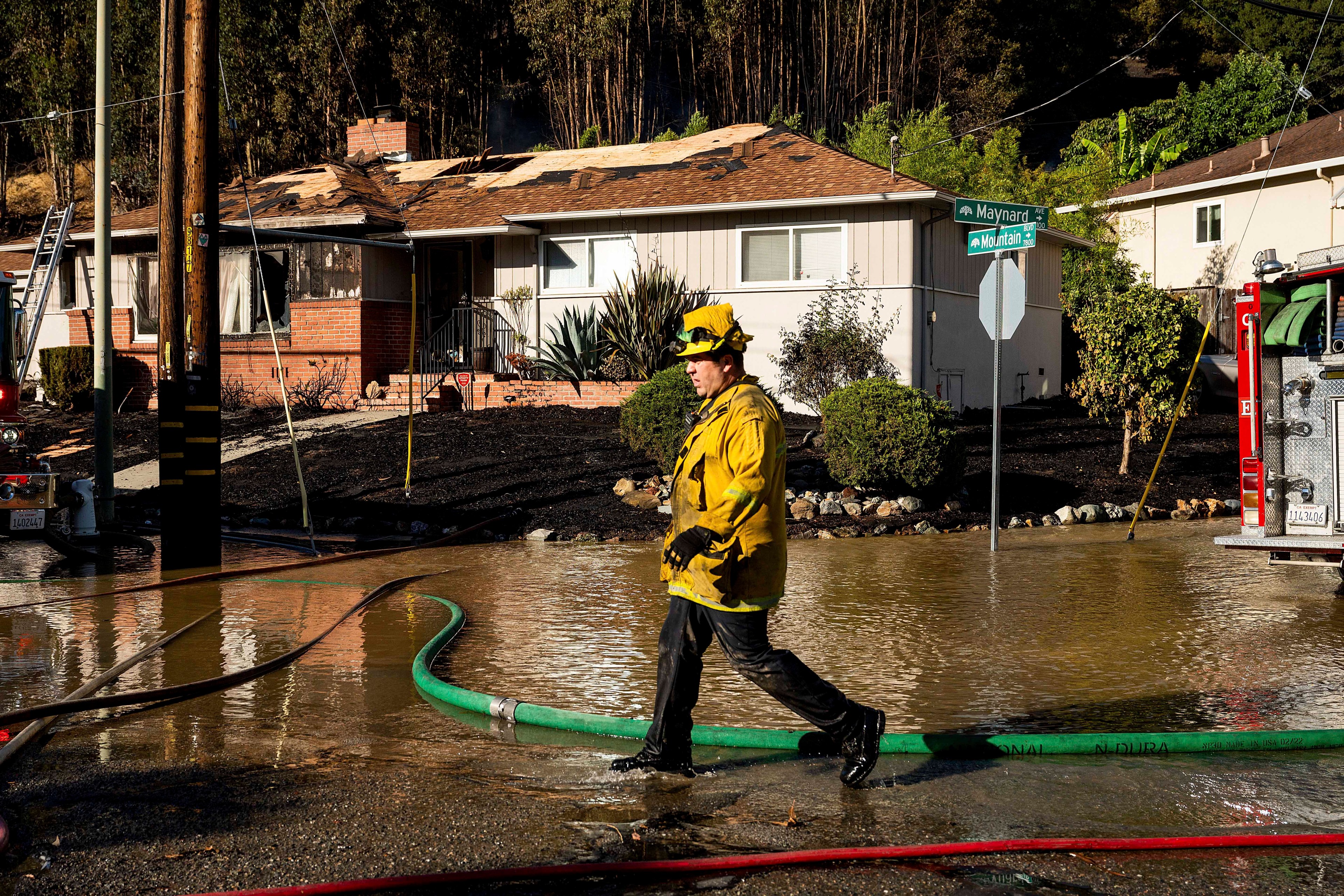 A firefighter in yellow gear walks through a flooded street, past a fire-damaged house with a charred roof, near a fire truck and a street sign.