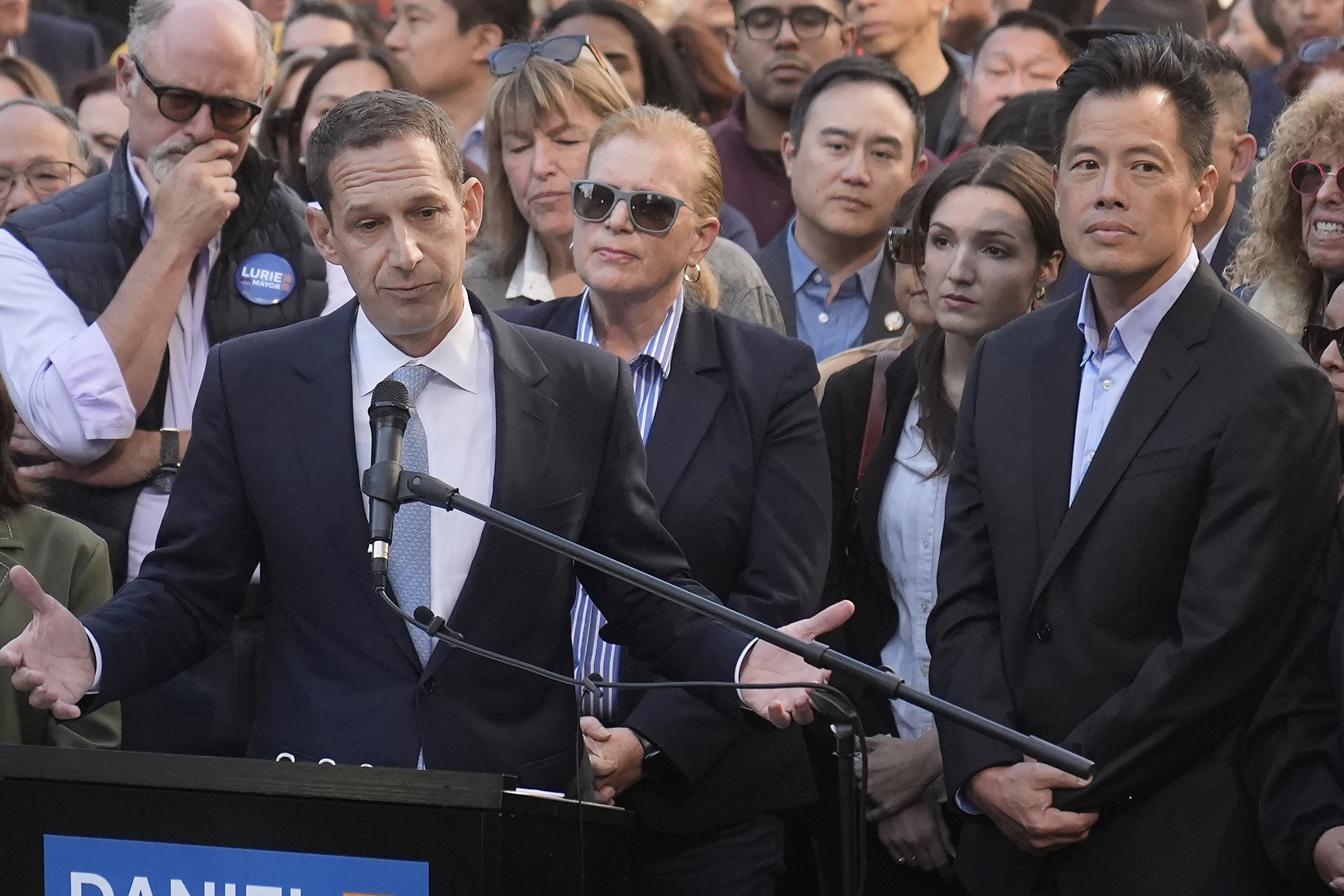 A man in a suit speaks at a podium with microphones, surrounded by a diverse crowd, including a woman in sunglasses and a man in a black suit.