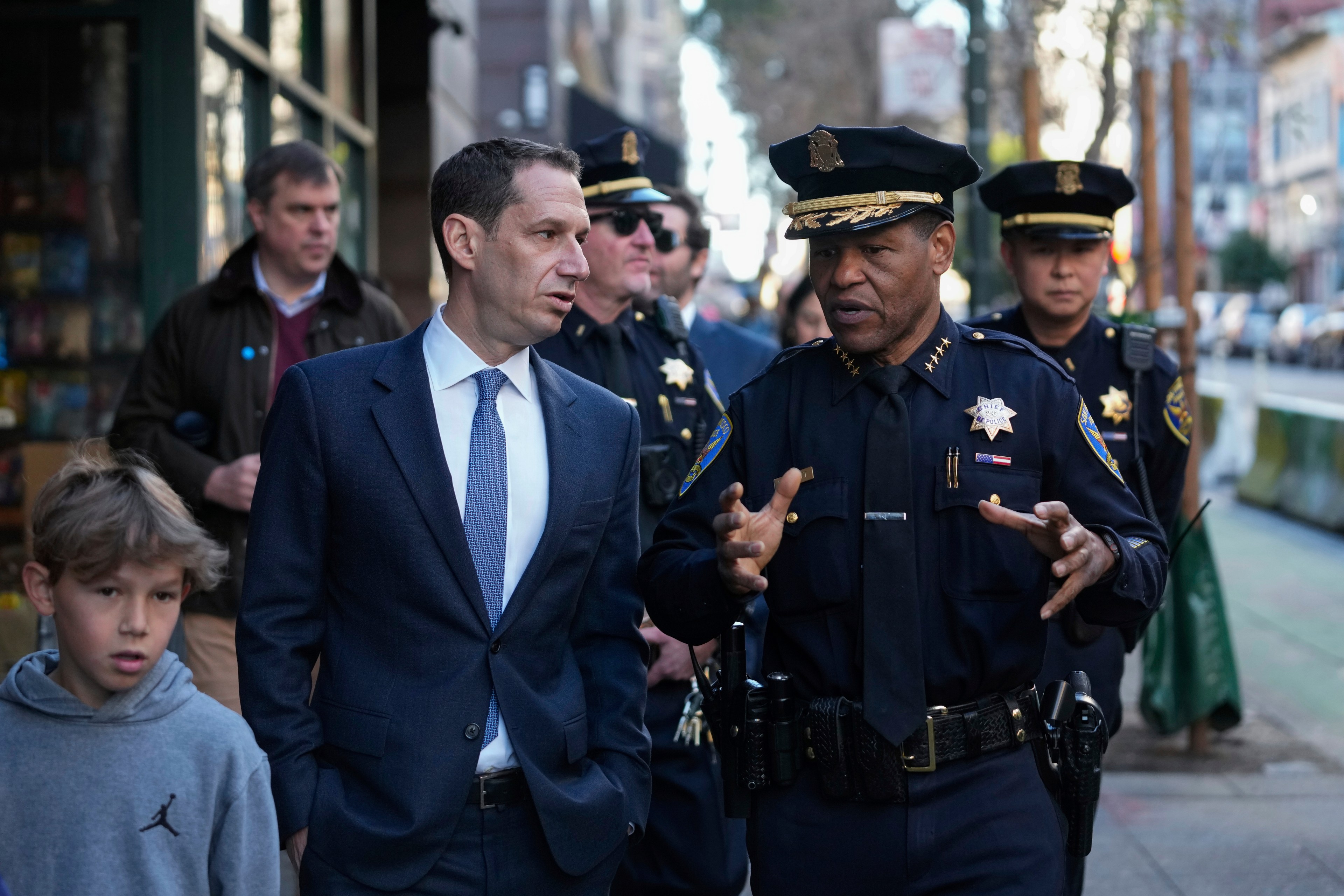 A police chief in uniform speaks with a man in a suit on a city street. Other officers and a young boy in a hoodie are nearby, with buildings in the background.