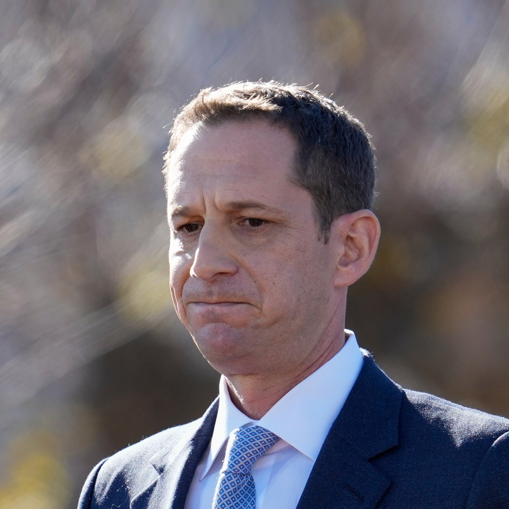 A man in a dark suit and light blue tie gazes pensively, set against a blurred outdoor background with soft lighting.