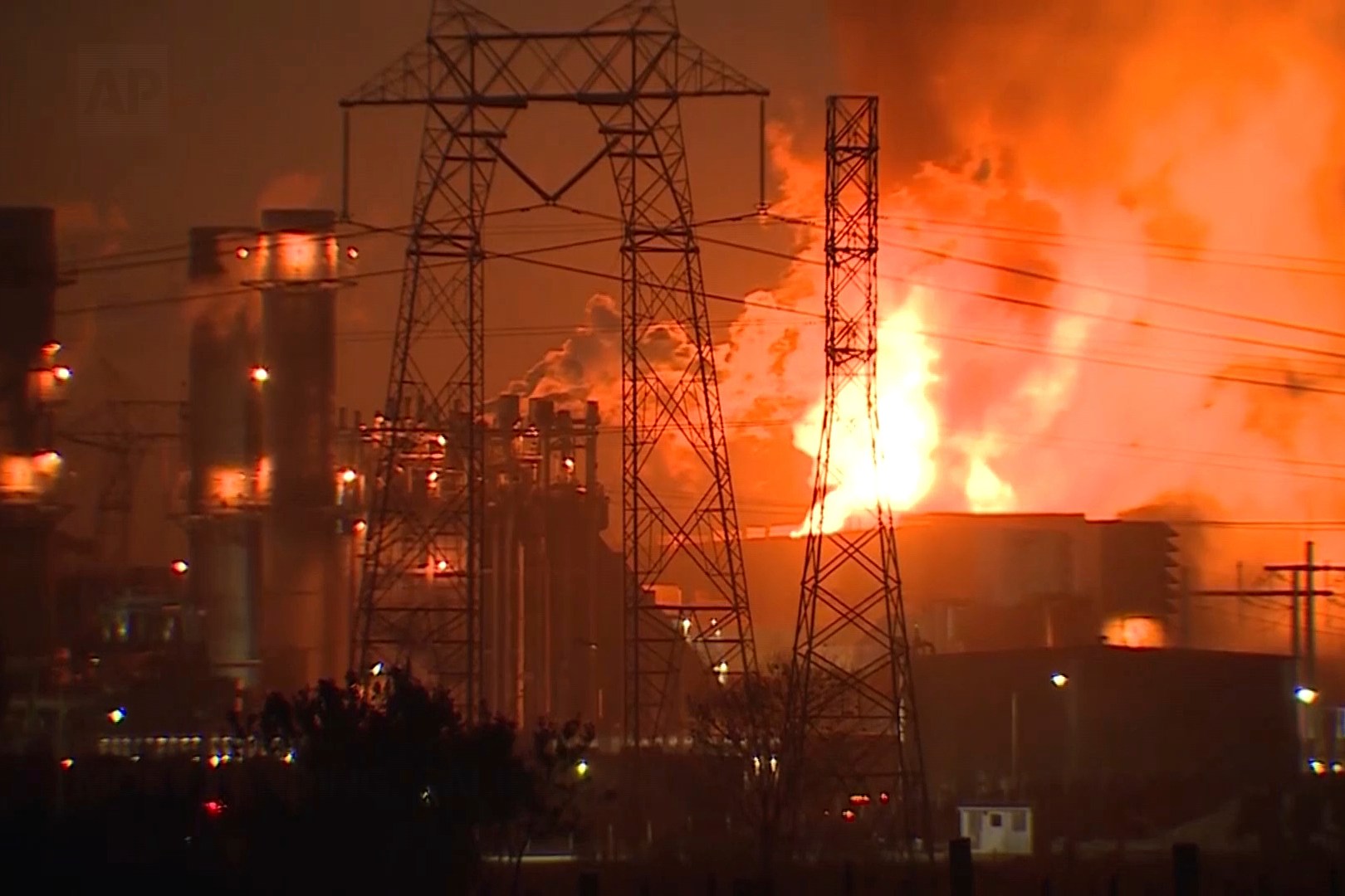 A large industrial complex is engulfed in flames, with bright orange and yellow fire illuminating the night. Power lines and structures are silhouetted.