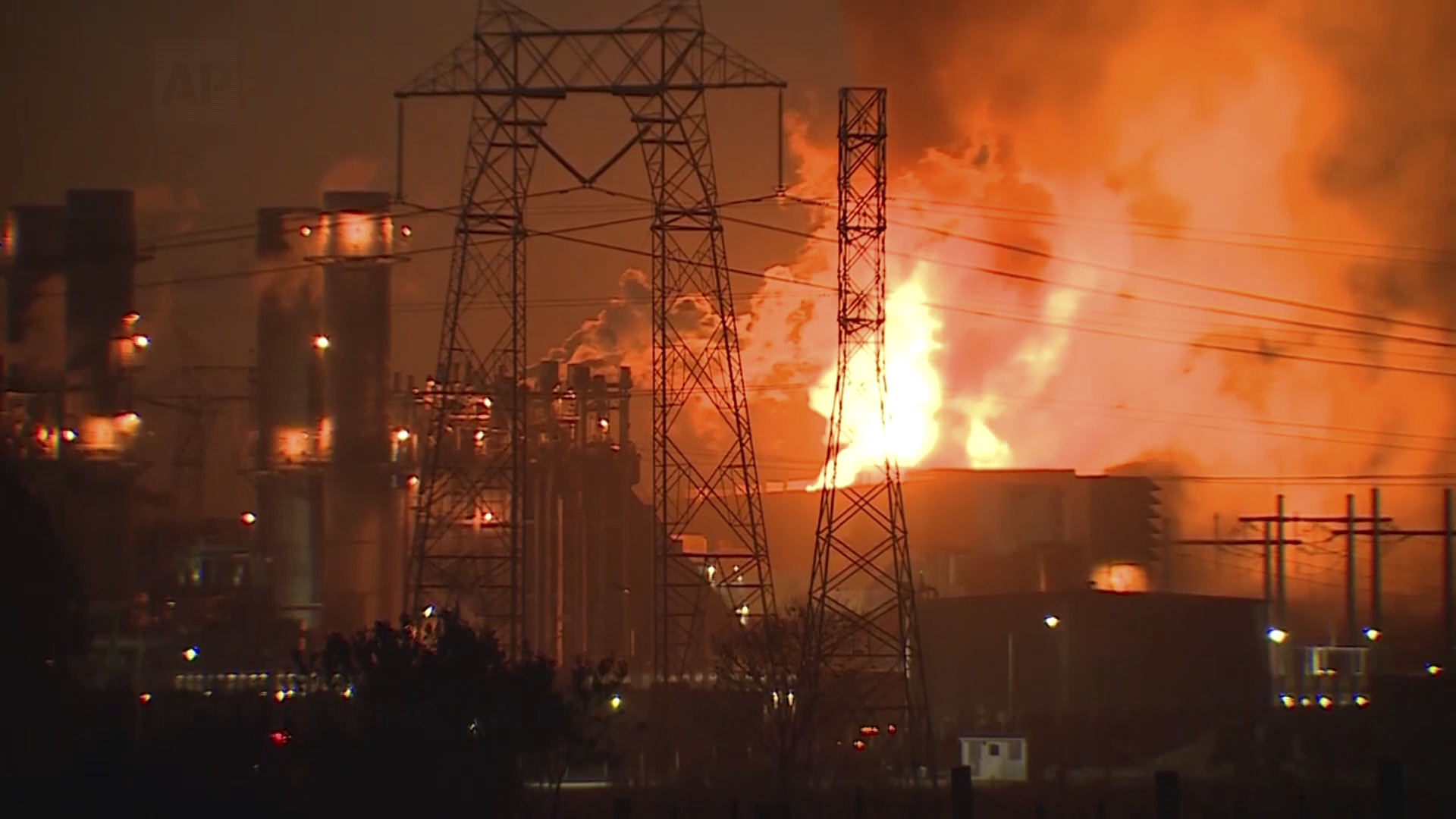 A large industrial complex is engulfed in flames, with bright orange and yellow fire illuminating the night. Power lines and structures are silhouetted.