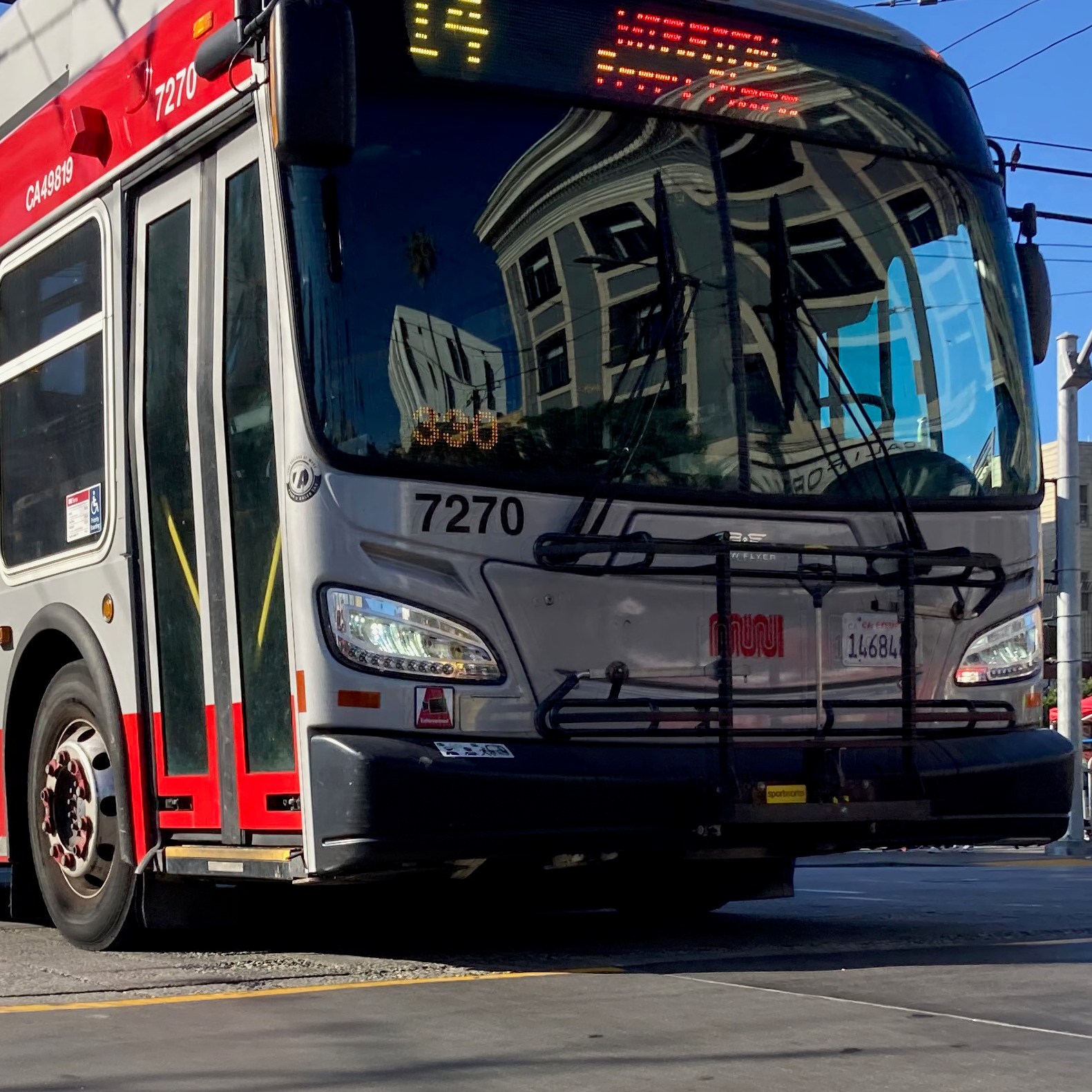 A red and gray city bus is driving on a street, with overhead wires visible above. It has the number 7270 on the front and a building reflection on its windshield.