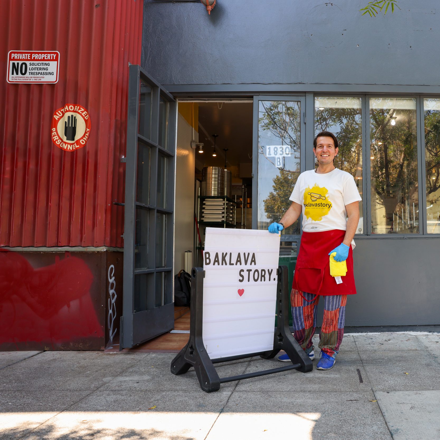 A person stands outside a store, smiling, wearing a white shirt and red apron. They're next to a sign that reads &quot;BAKLAVA STORY.&quot; The store has a red wall.