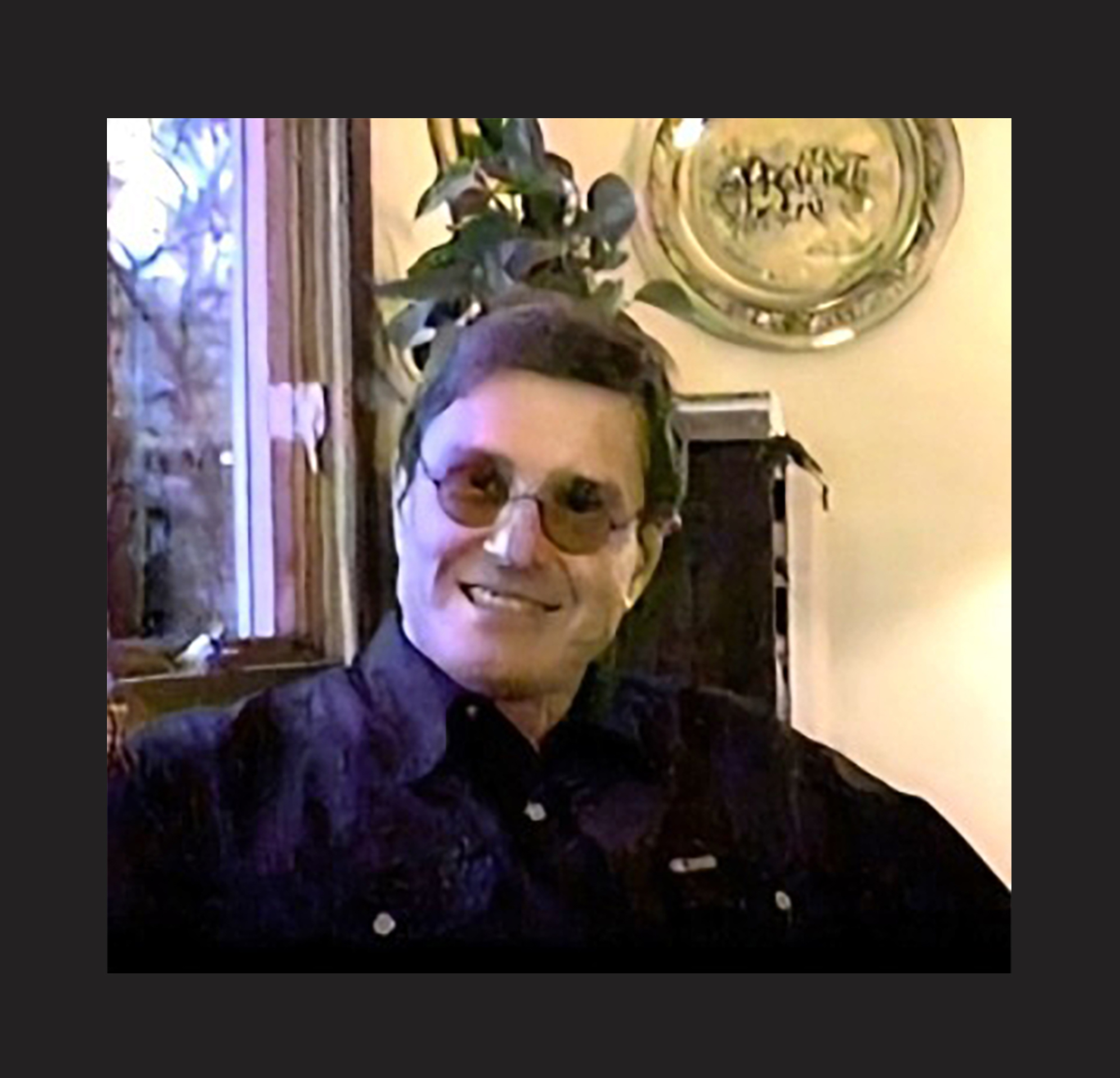 A man wearing sunglasses and a dark shirt smiles while seated indoors. There is a window, a plant, and a decorative plate on the wall behind him.